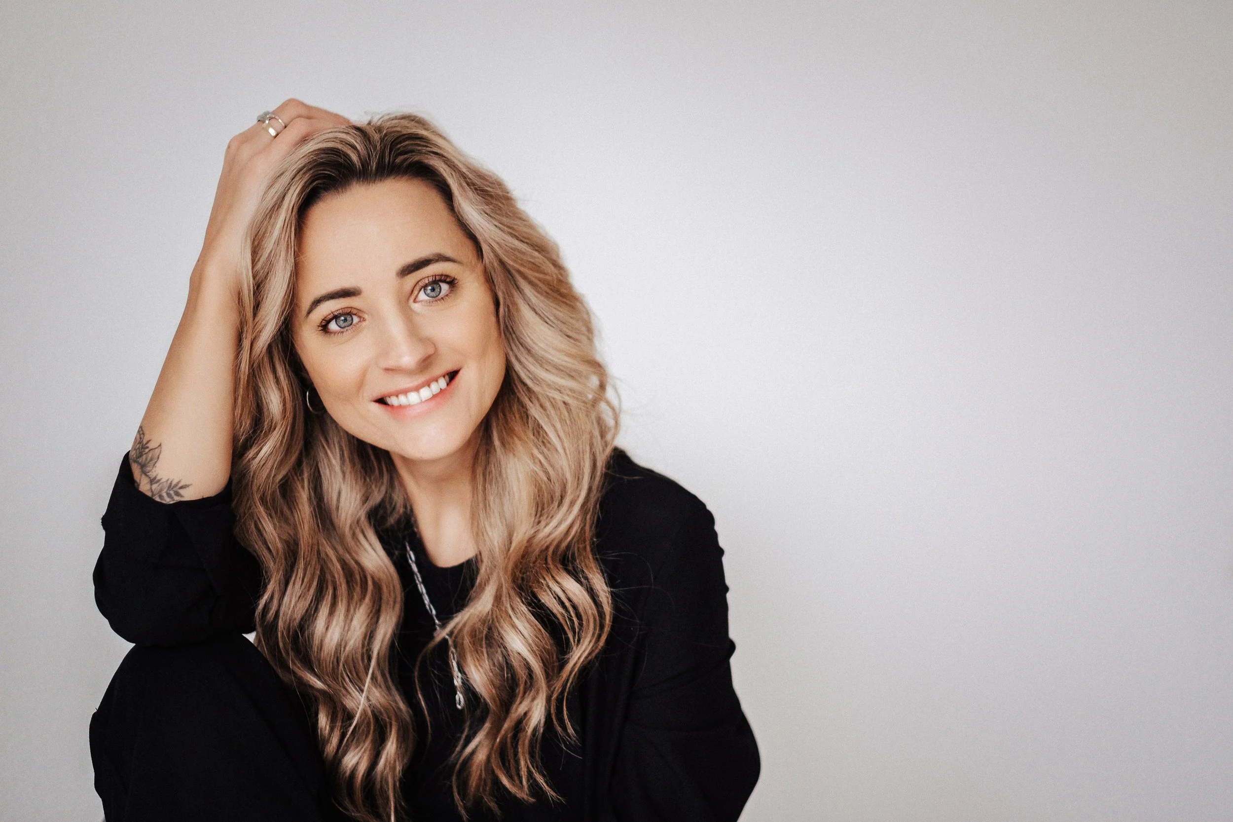 Lächelnde Frau mit langen, gewellten Haaren, die eine Hand auf dem Kopf hat, trägt schwarze Kleidung vor einem neutralen Hintergrund.