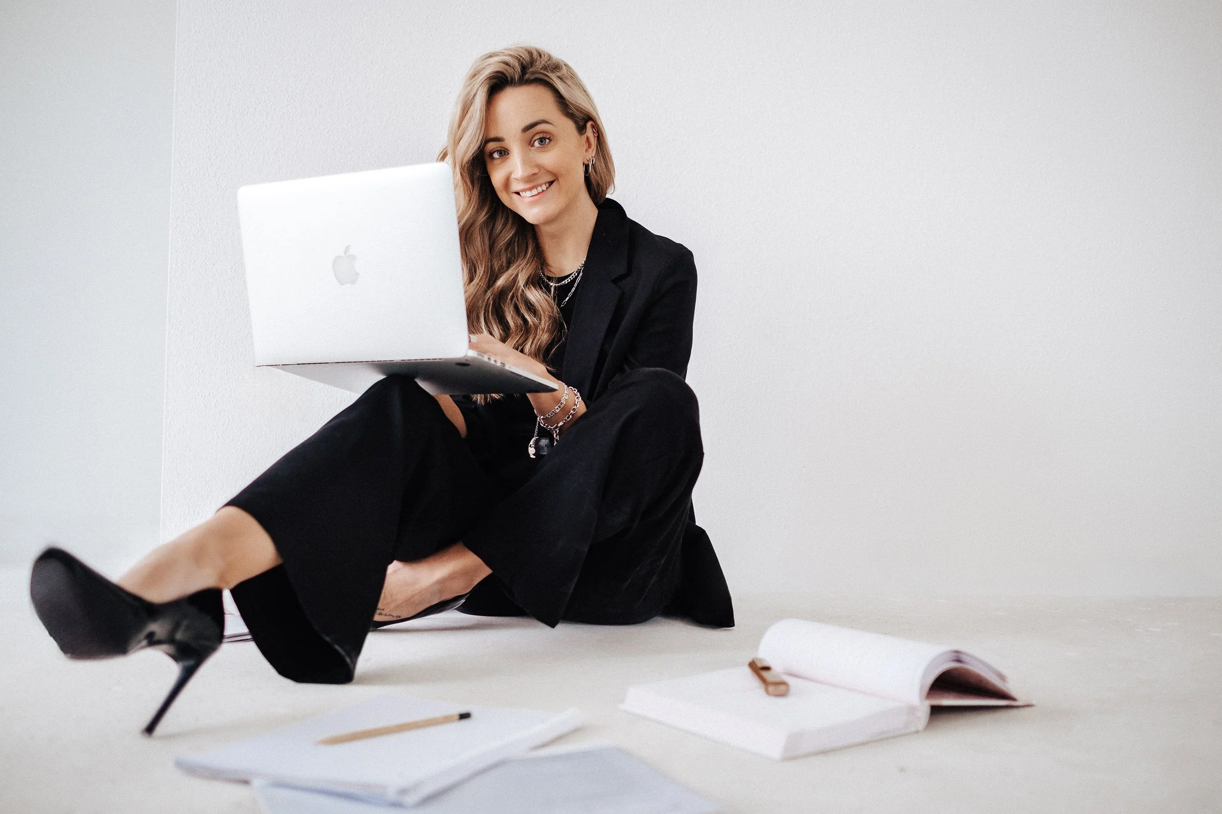 Junge Frau mit langen, gewellten Haaren sitzt auf dem Boden, hält einen MacBook-Laptop, trägt schwarze Kleidung und High Heels, umgeben von offenen Notizbüchern und Schreibmaterialien.