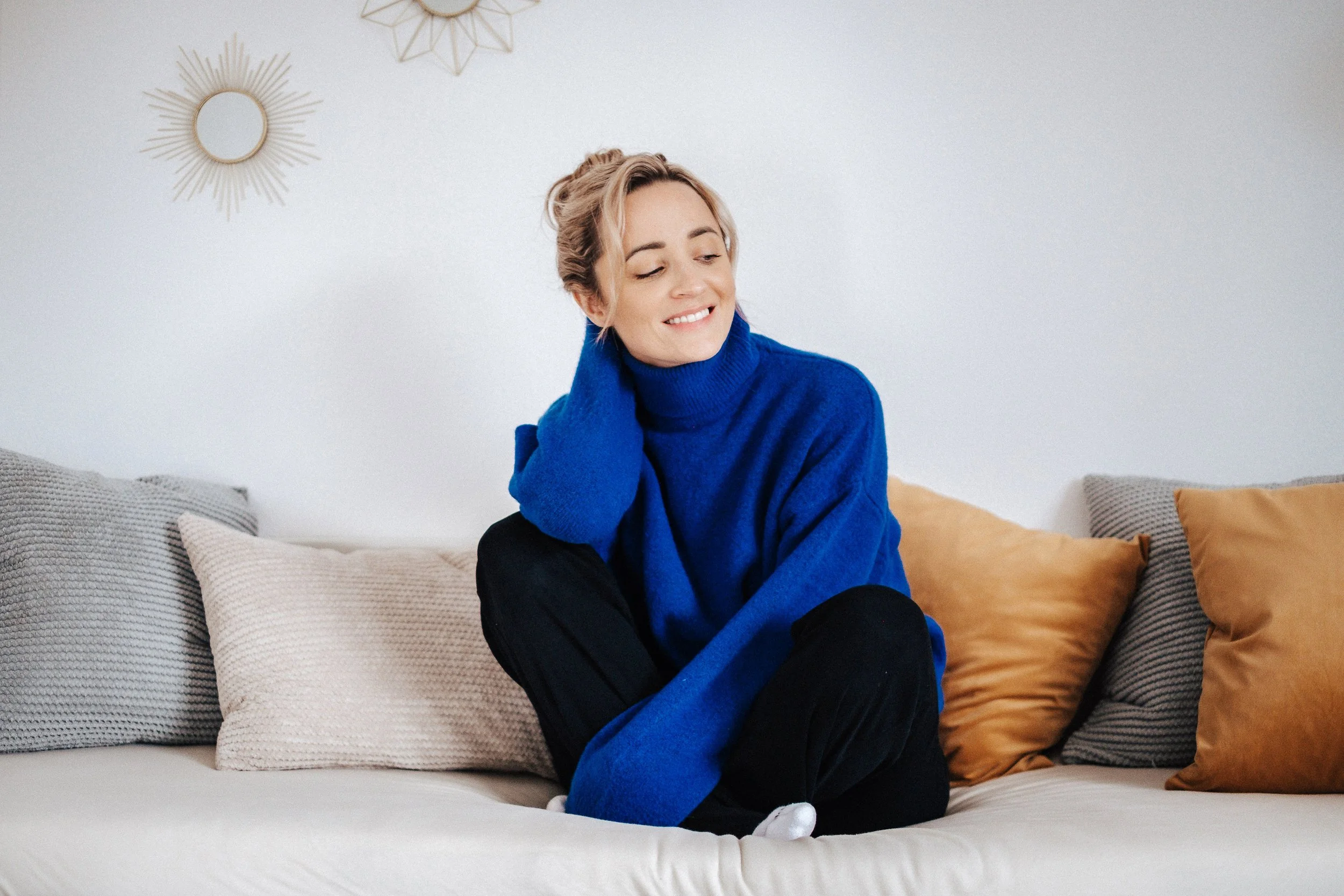 Junge Frau sitzt entspannt auf einem Sofa mit bunten Kissen, lächelt und trägt einen blauen Pullover.