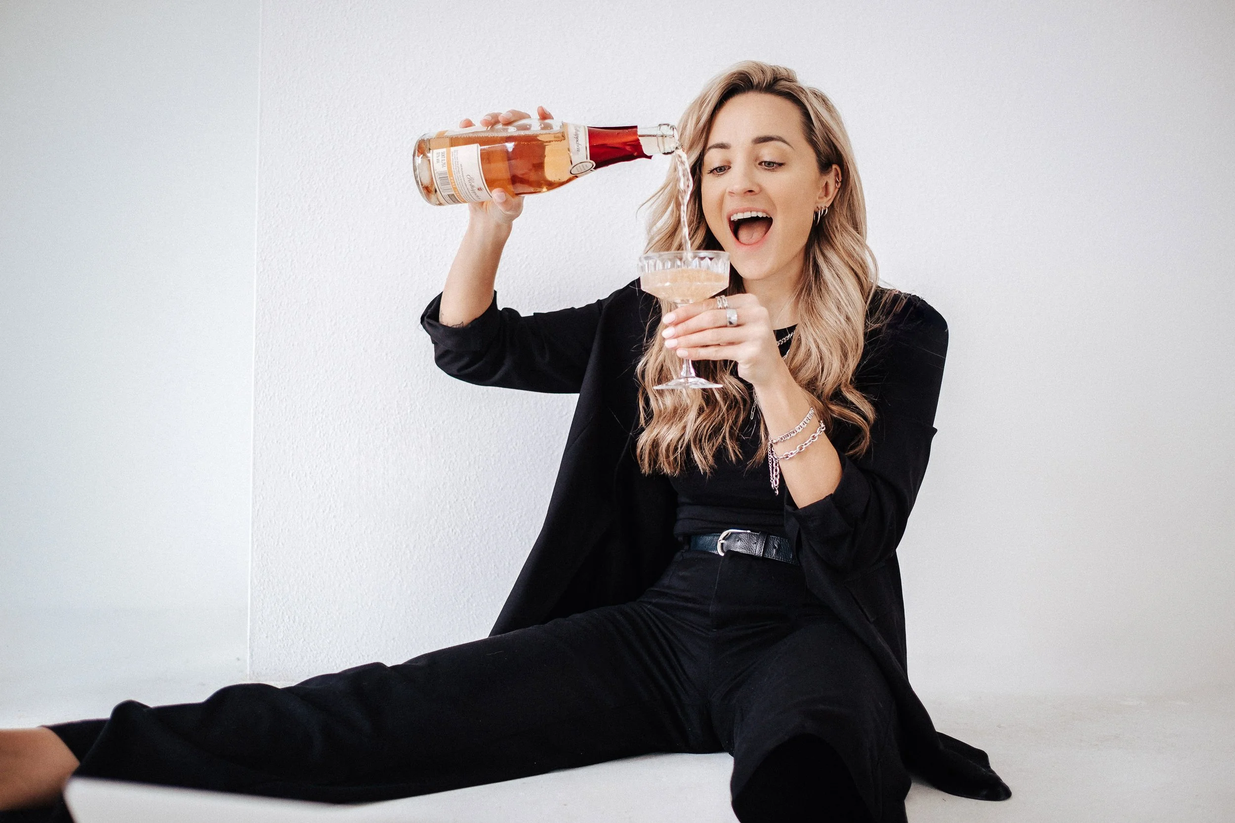 Eine junge Frau sitzt auf dem Boden und schenkt sich Sekt ein, während sie eine Flasche Sekt hält, den sie gerade eingießt, in einem minimalistischen weißen Raum.