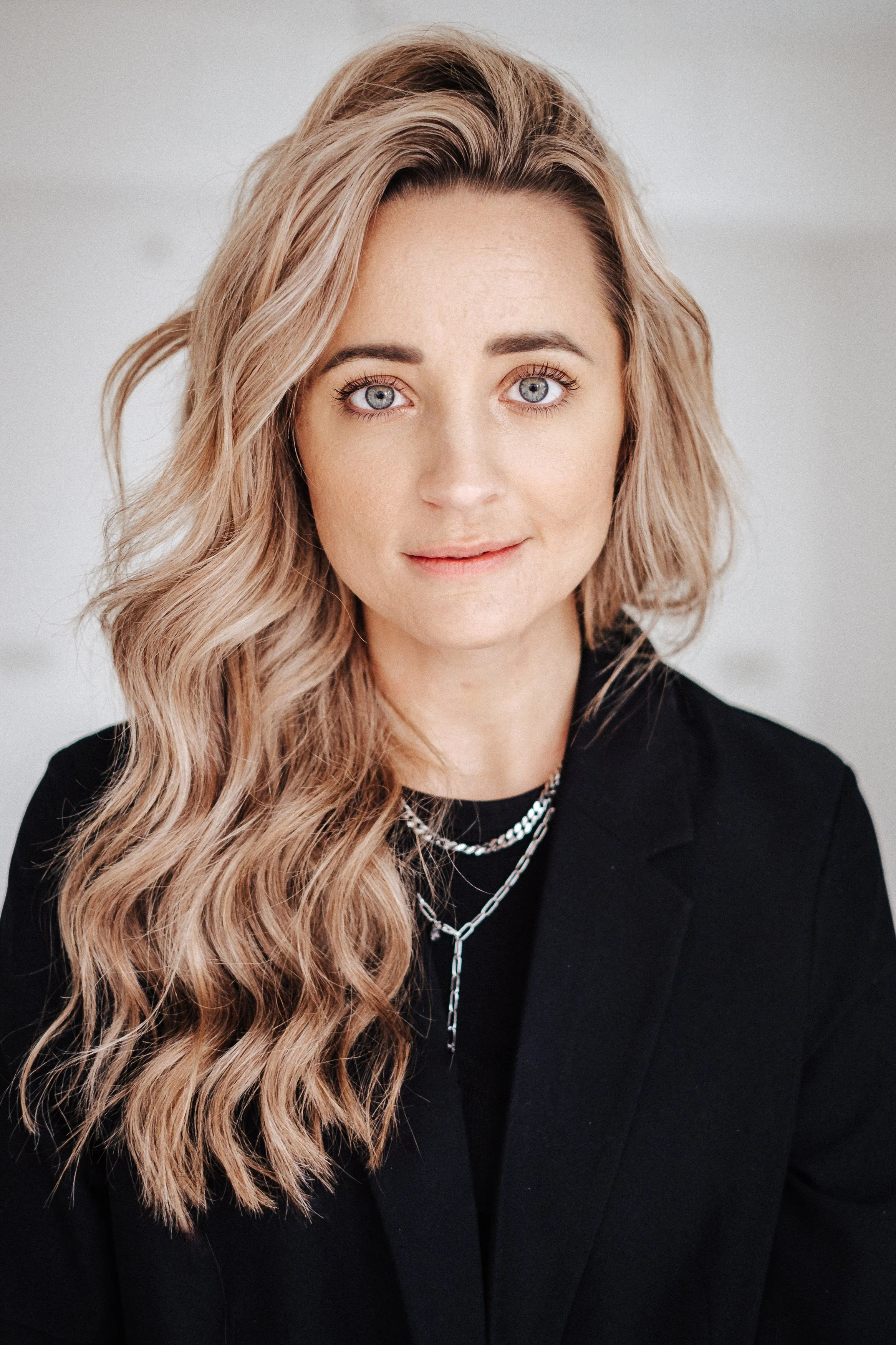 Porträt einer Frau mit blonden, lockigen Haaren, blauen Augen und einem schwarzen Blazer, trägt mehrere silberne Halsketten.