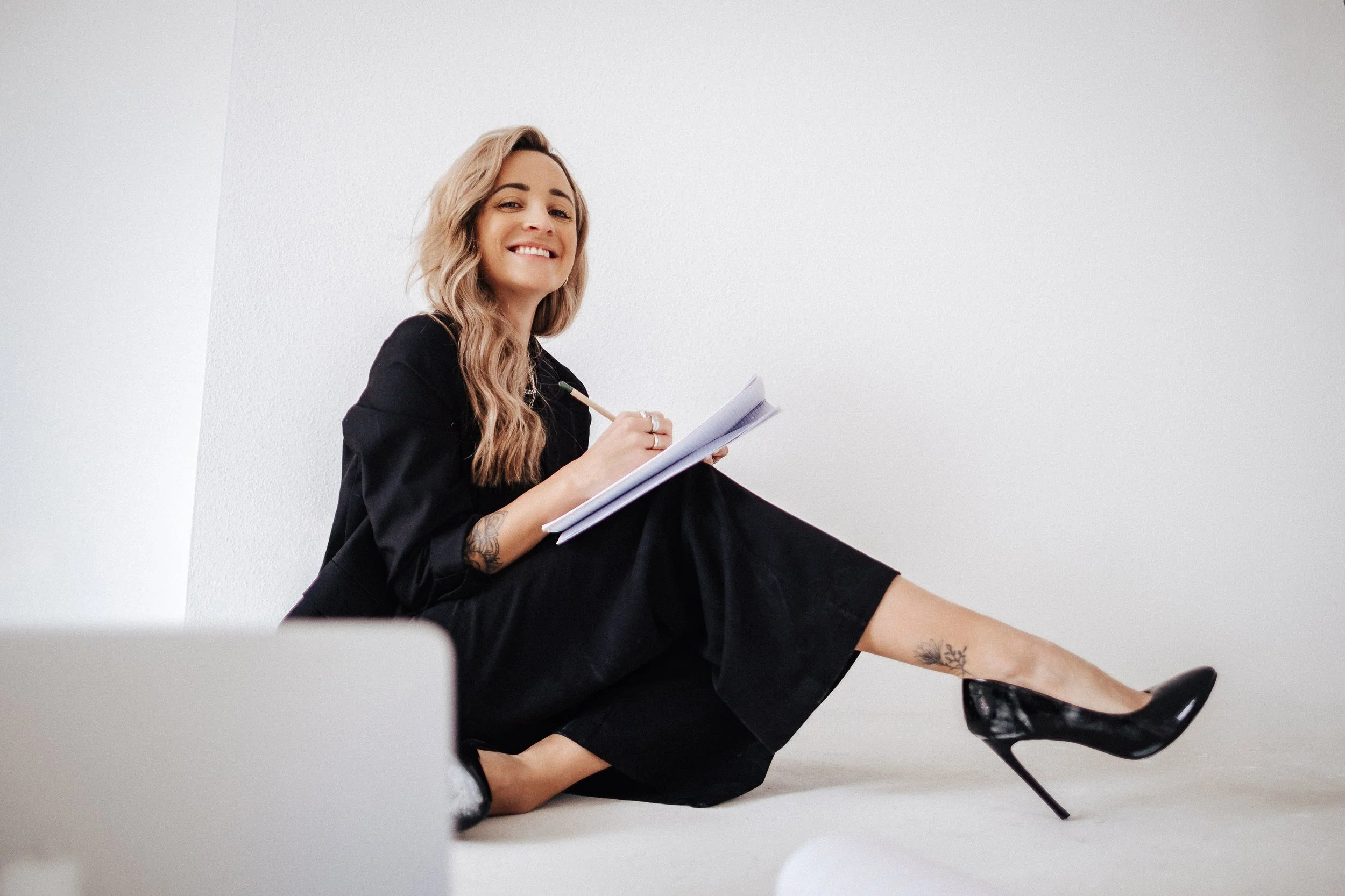 Junge Frau sitzt auf dem Boden, trägt schwarze Kleidung und High Heels, schreibt in ein Notizbuch und lächelt.