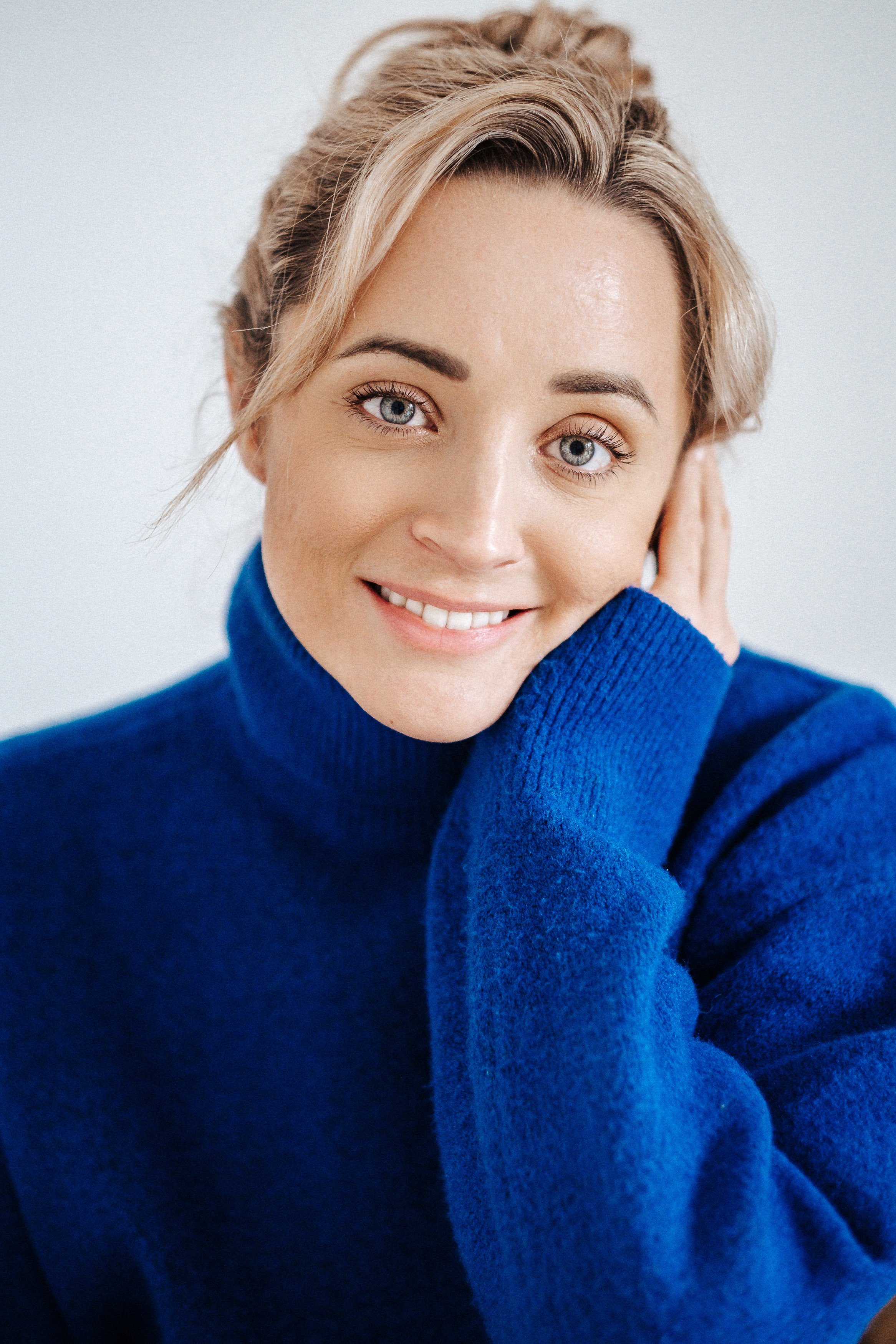 Nahaufnahme einer lachenden Frau mit blonden Haaren und blauen Augen, trägt einen blauen Pullover, die Hand berührt ihr Ohr.