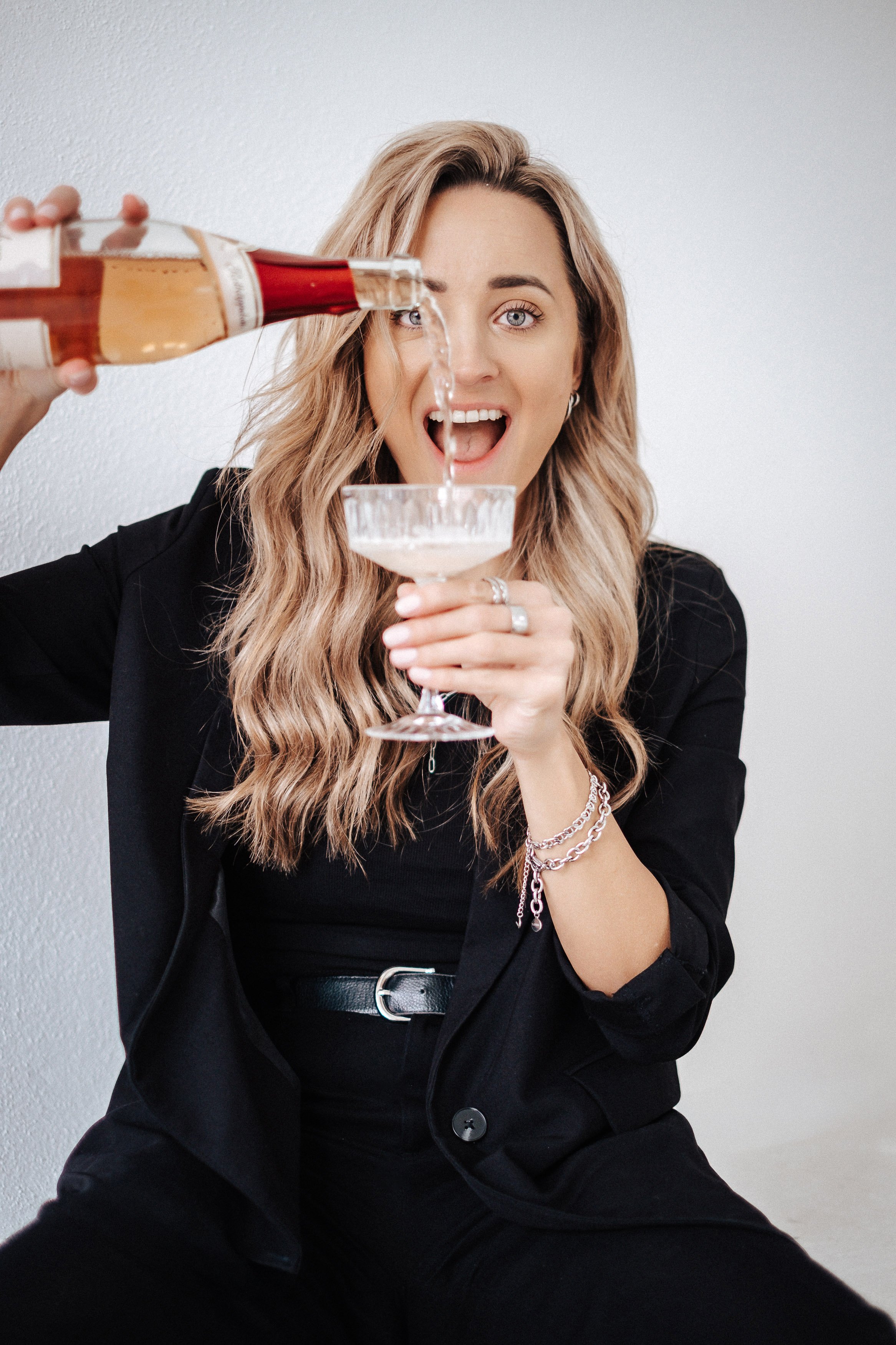 Eine Frau in schwarzer Kleidung, die mit einem Cocktailglas in der Hand sitzt, während jemand mit einer Flasche Sekt darauf einschenkt.