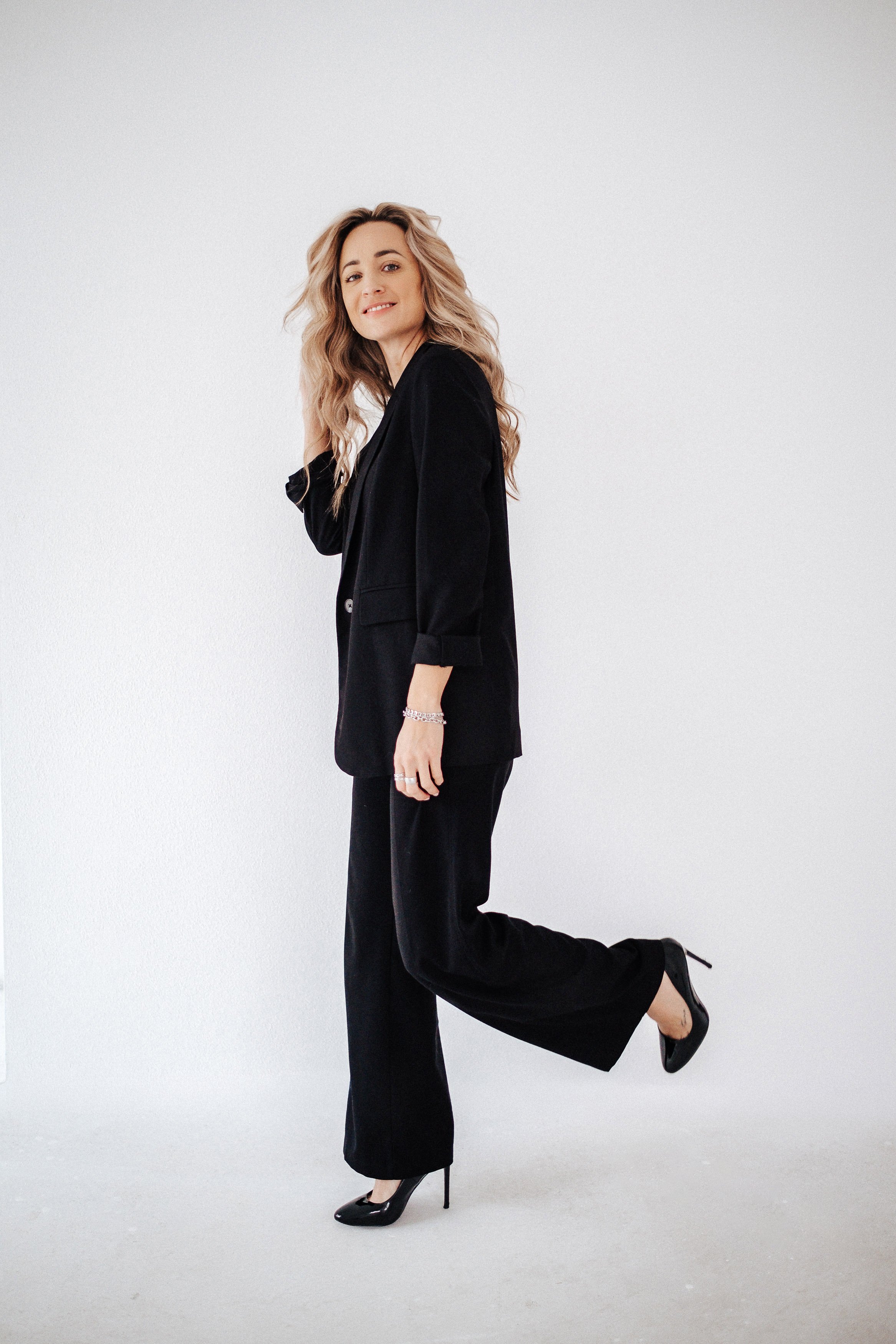 Eine Frau in einem schwarzen Anzug und hohen Schuhen posiert vor einem weißen Hintergrund.
