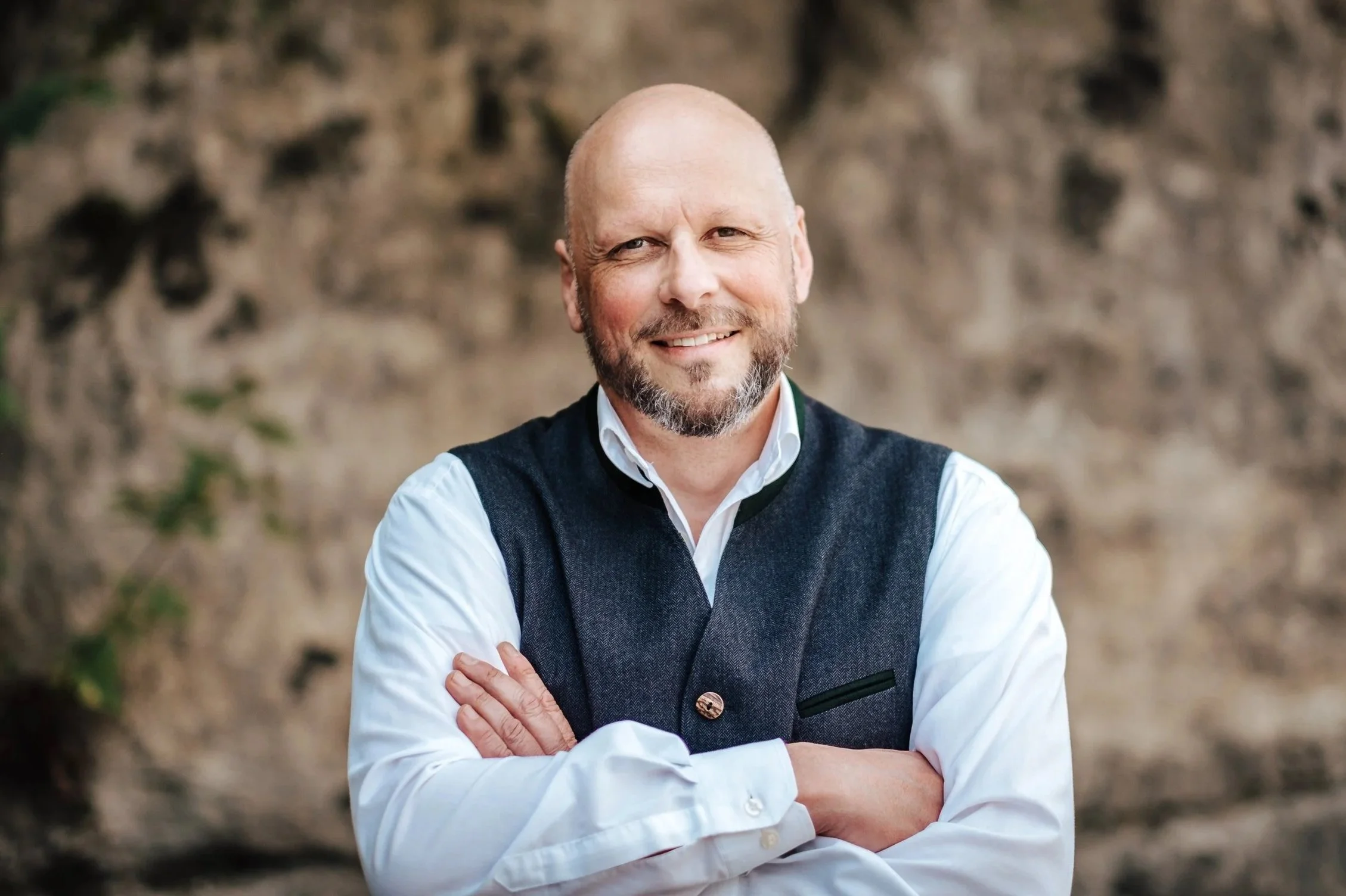 Ein lächelnder Mann mit Bart, trägt ein weißes Hemd und eine dunkle Weste, steht vor einer natürlichen Stein- und Pflanzenhintergrund.
