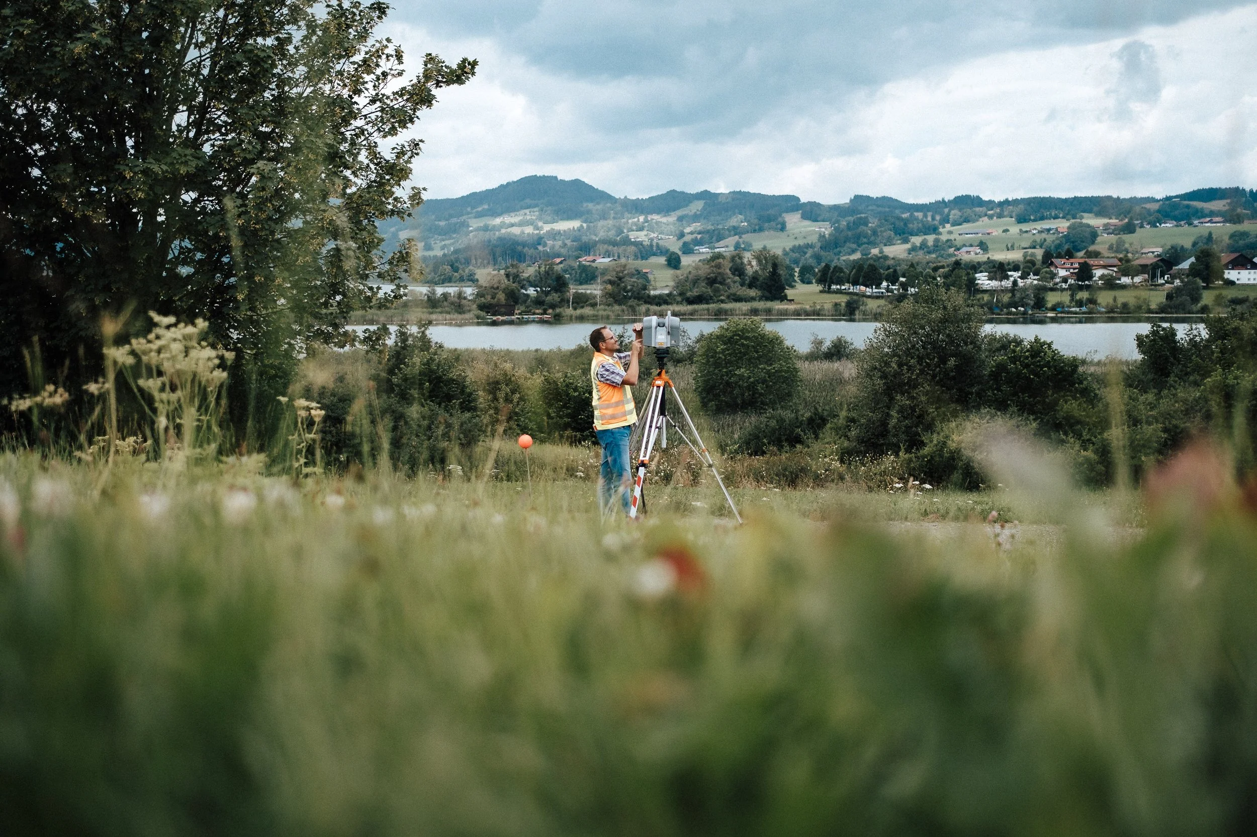 Ein Mann steht mit einer Vermessungsausrüstung auf einer Wiese in einer ländlichen Gegend, mit einem See und Hügel im Hintergrund.