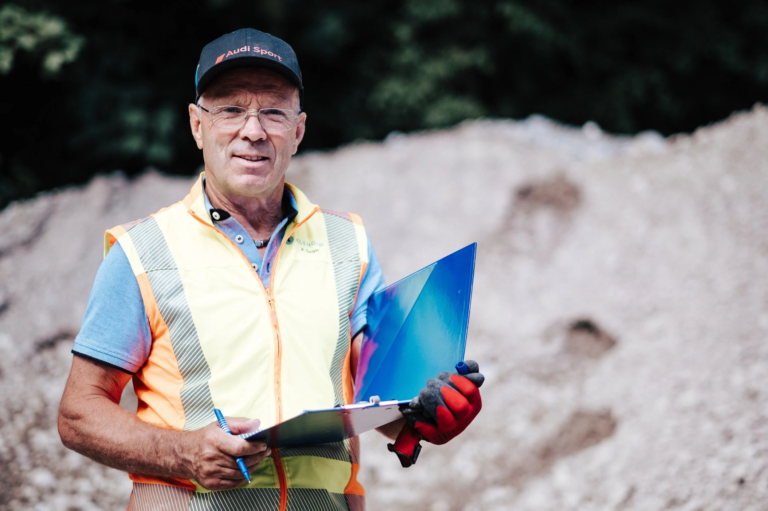 Ein älterer Mann mit Brille, schwarzem Cap mit rotem Schriftzug, gelbe Sicherheitsweste, blauer Kurzarmshirt und roten Arbeitshandschuhen steht vor einem Sandhaufen und hält eine blaue Aktenmappe und einen Kugelschreiber. Er schaut in die Kamera.