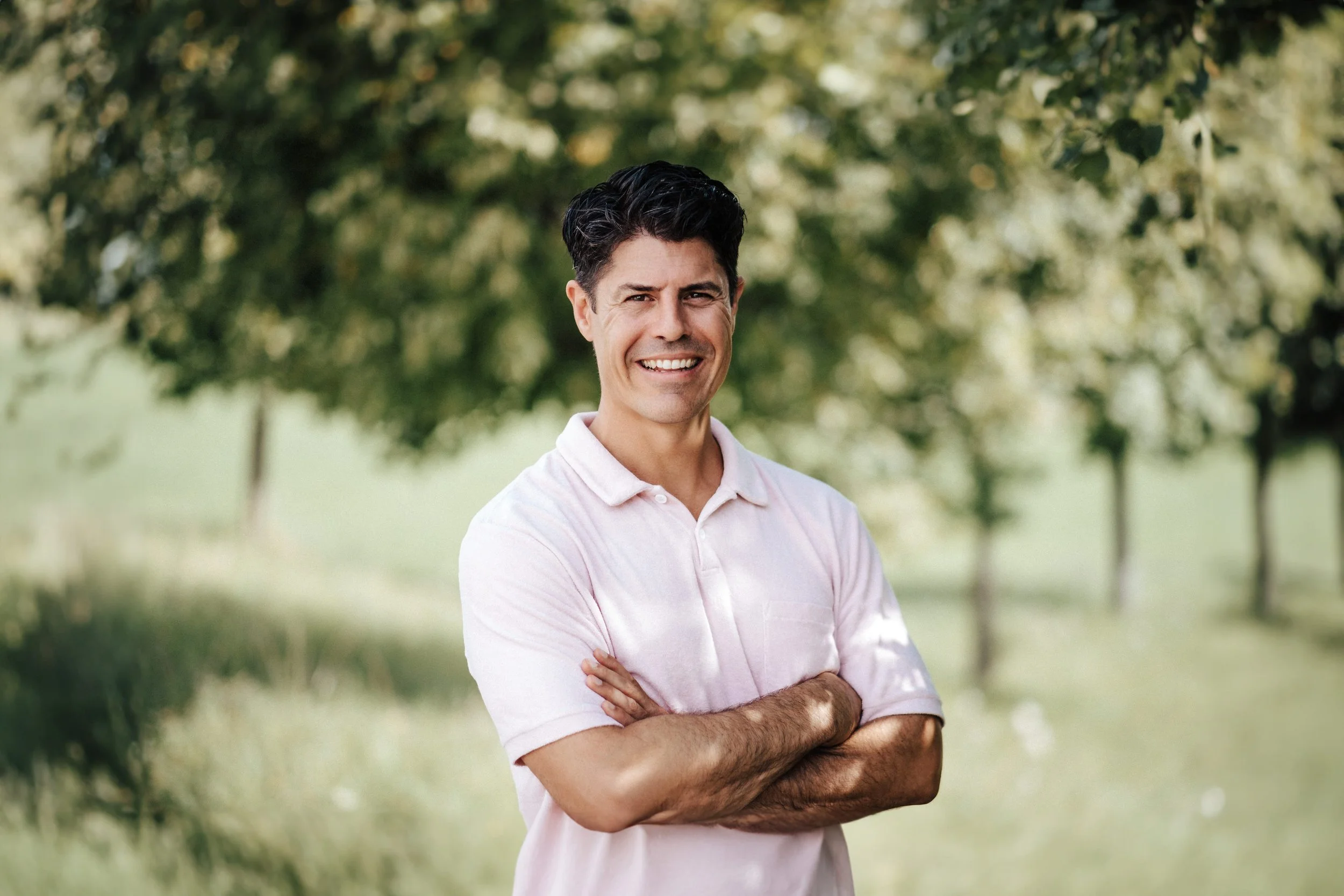 Ein Mann mit dunklem Haar trägt ein hellrosa Polohemd, steht in einem Park mit grünen Bäumen und lächelt in die Kamera.