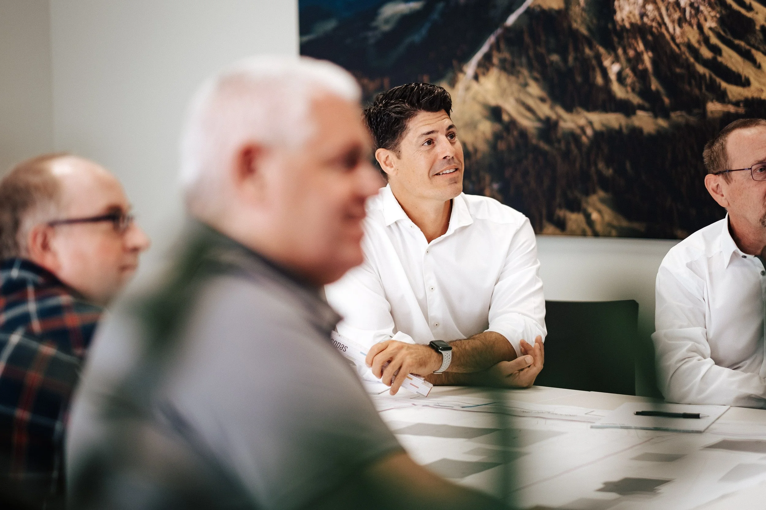 Vier Männer sitzen in einem Meetingraum, der Mann in der Mitte trägt ein weißes Hemd und schaut aufmerksam. Hinter ihm eine Fototapete mit einer Berglandschaft. Die anderen Männer sind unscharf im Vordergrund, es herrscht eine konzentrierte Atmosphär