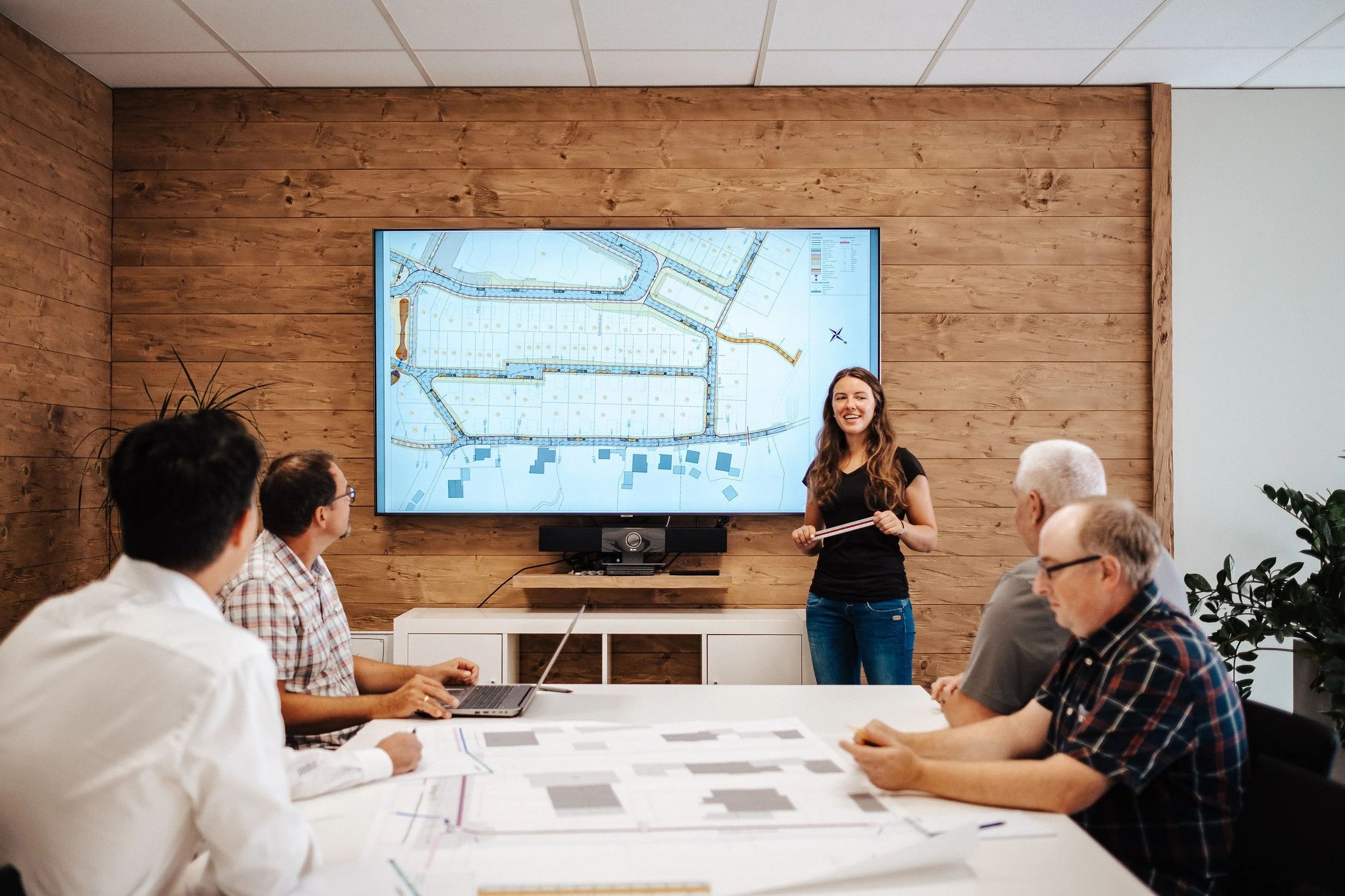Frau präsentiert einen Stadtplan an einer TV-Wand in einem Konferenzraum, während fünf Personen dem Vortrag zuhören.