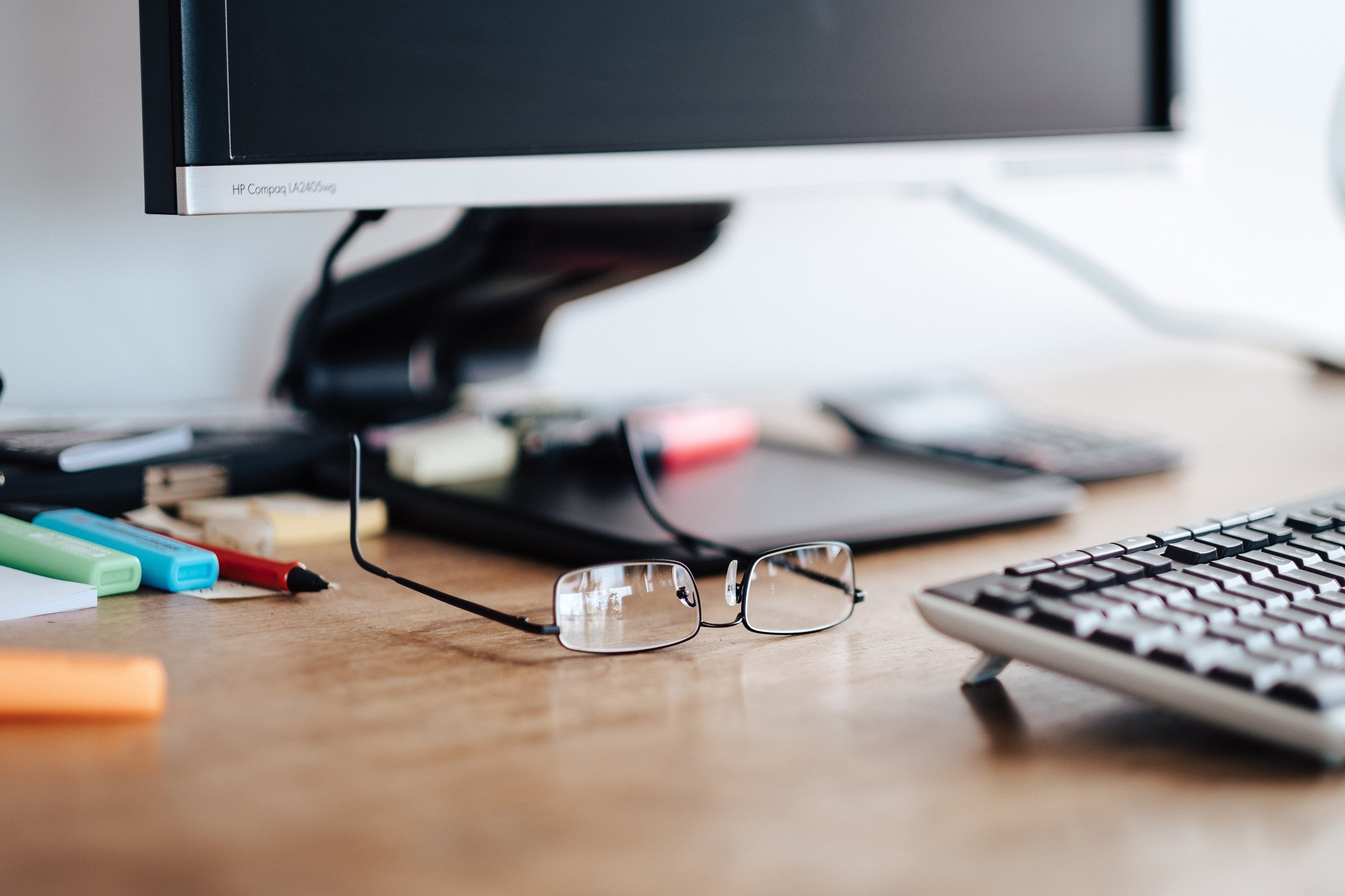 Arbeitsplatz mit Brille, Stiften, Tastatur, Bildschirm und anderen Gegenständen auf einem Holztisch.