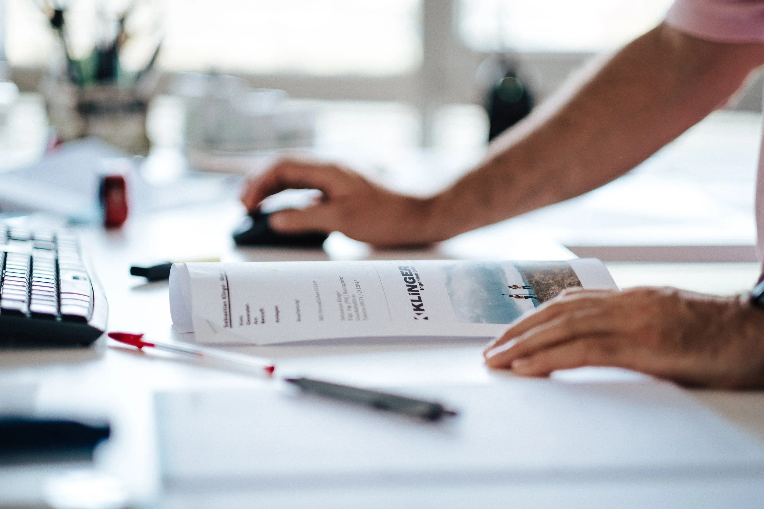 Ein Mensch arbeitet an einem Schreibtisch mit einem Computer-Tastatur, einer Zeitschrift und einem Stift.