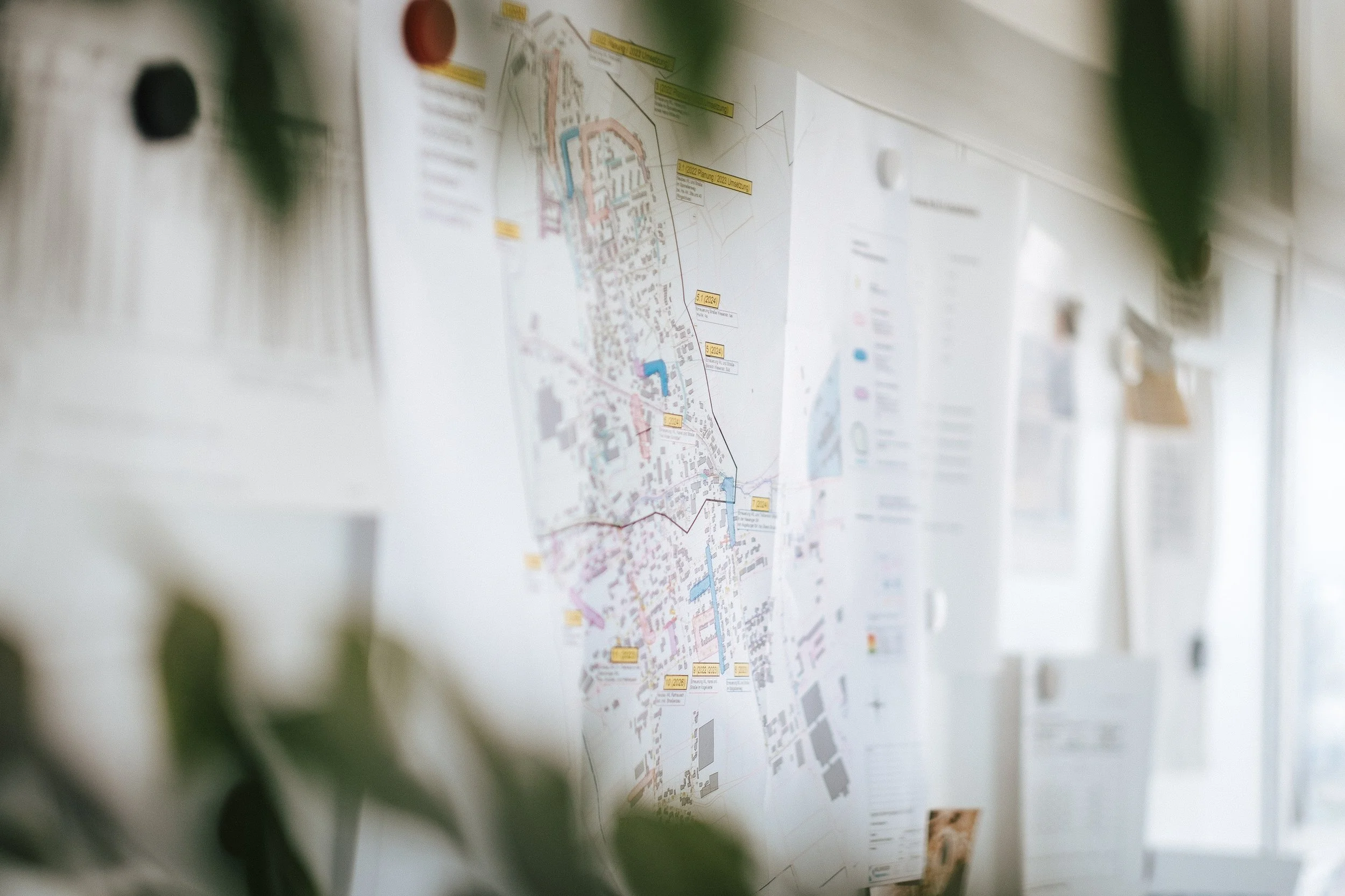 Karte oder Plan an einer Wand, teilweise sichtbar, mit verschiedenen Markierungen und Texten. Im Vordergrund und auf der Seite sind unscharfe grüne Blätter.