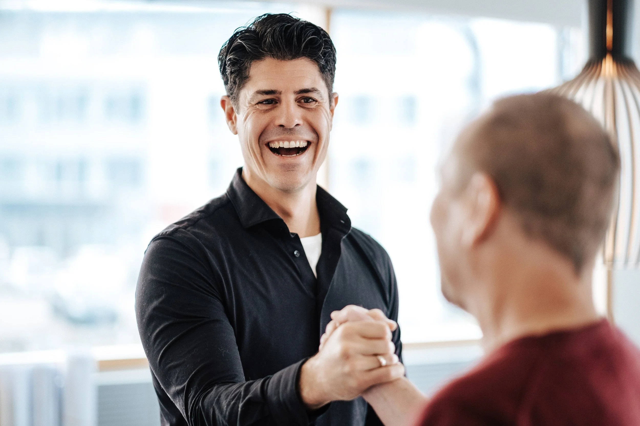 Zwei Männer schütteln sich die Hände und lachen. Der Mann im Vordergrund trägt ein schwarzes Hemd, der andere trägt eine dunkelrote Jacke. Sie scheinen sich zu begrüßen oder zu freuen.