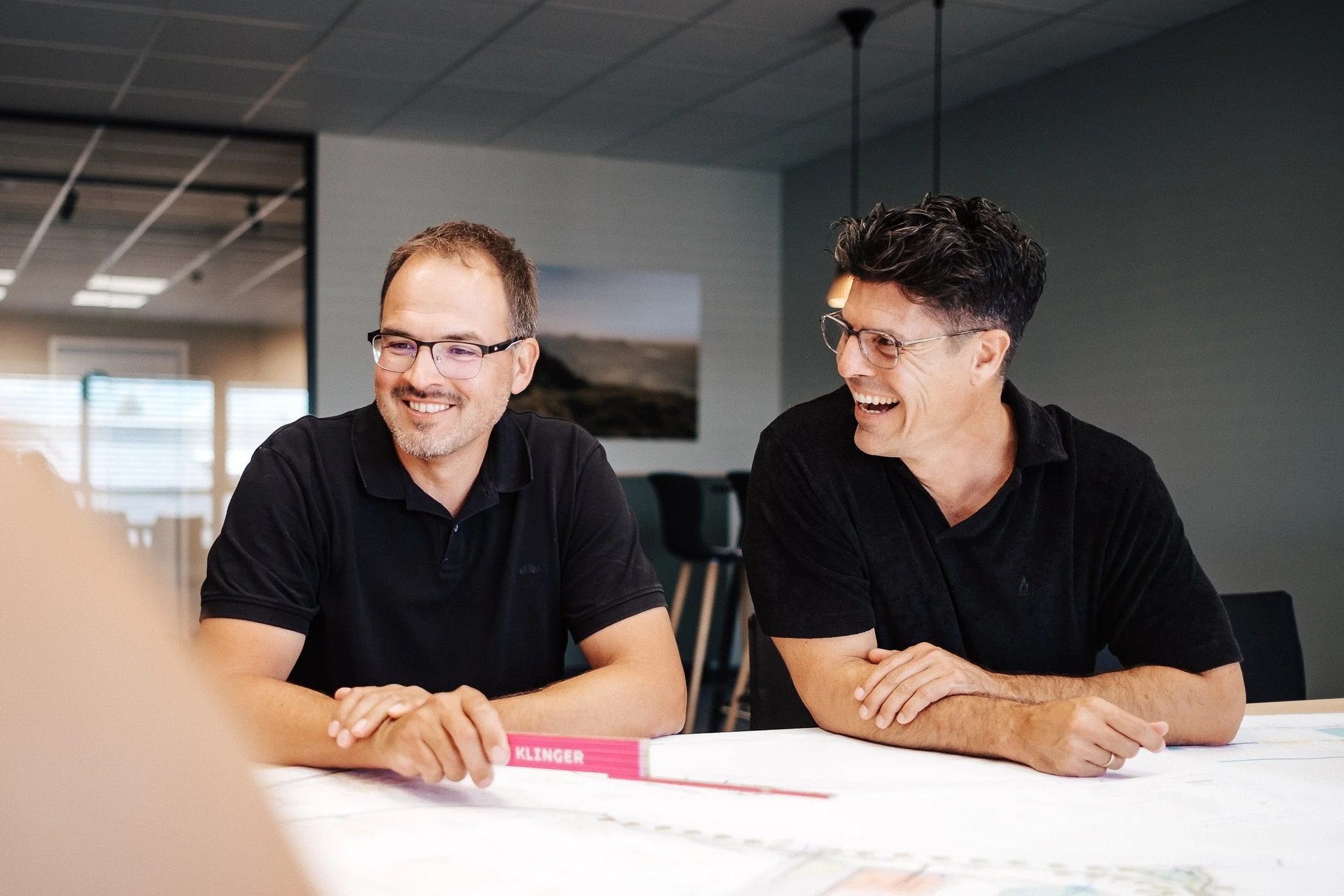 Zwei lachende Männer in schwarzen T-Shirts sitzen an einem Tisch und unterhalten sich in einem modernen Büroraum.