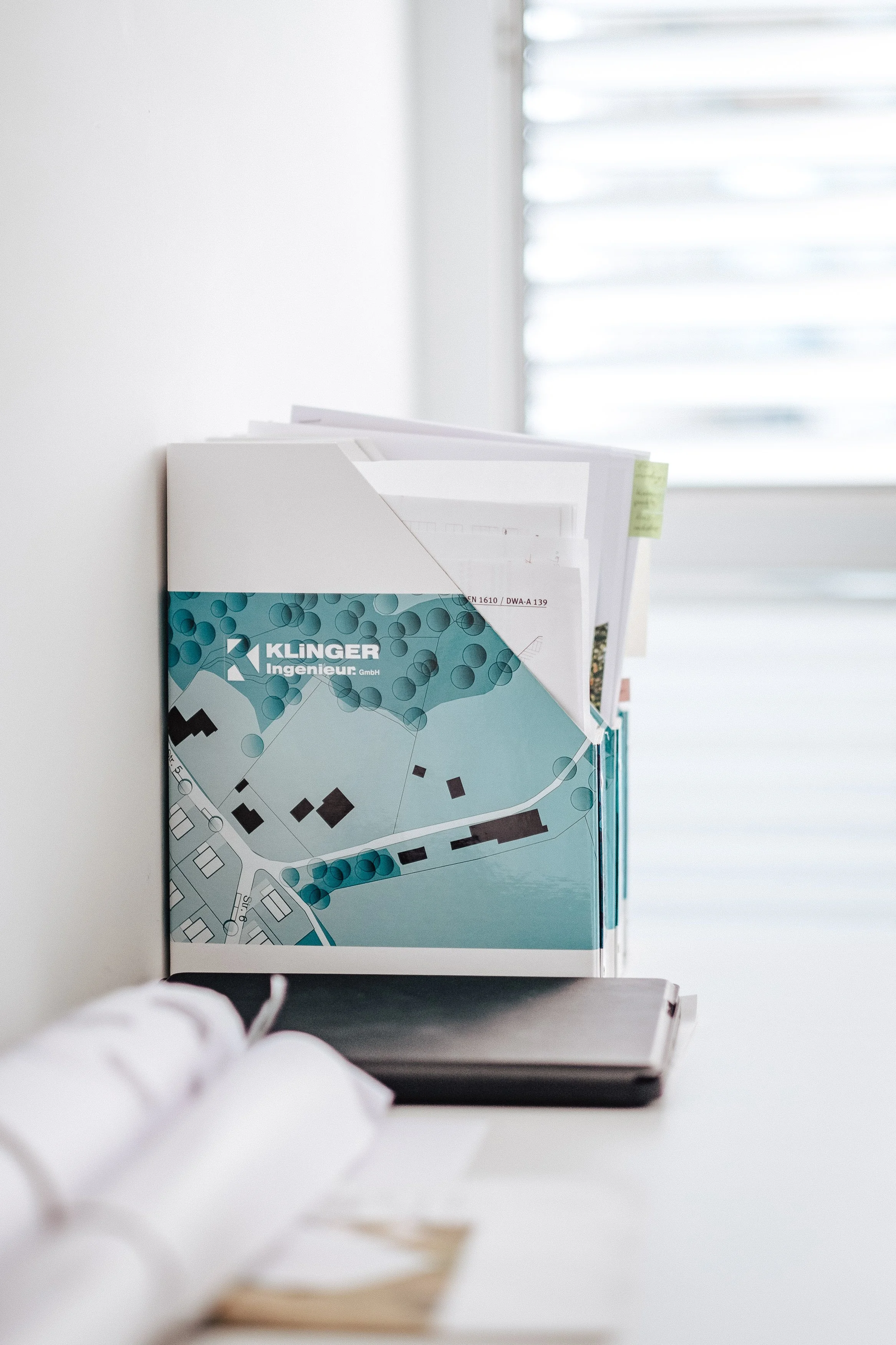 Büroablage mit technischen Unterlagen und einem Mappeinhalt mit der Aufschrift "Klinger Ingenieur GmbH" auf einem weißen Schreibtisch, im Hintergrund ein Fenster mit geschlossenen Jalousien.