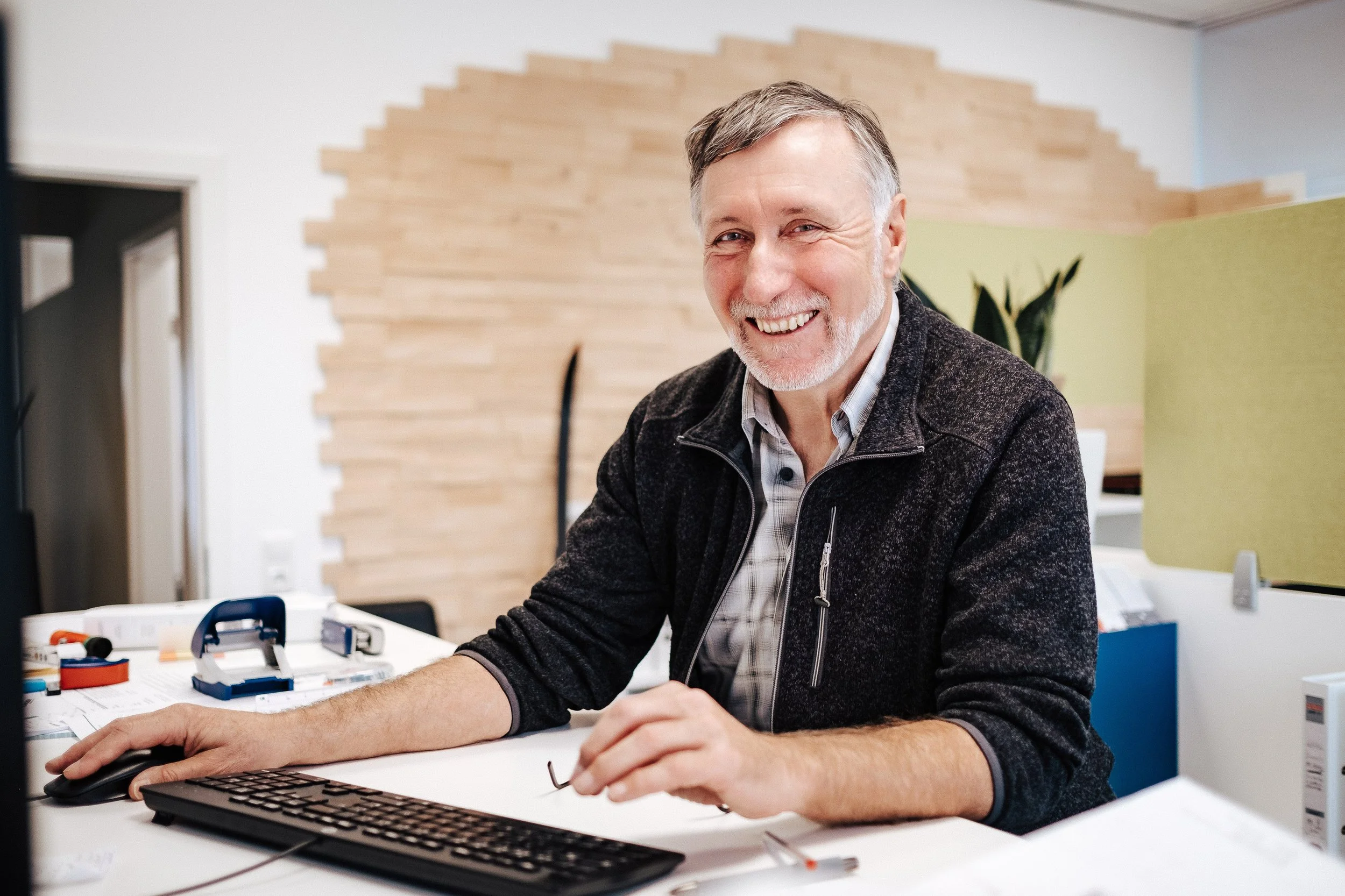 Lächelnder älterer Mann sitzt am Arbeitsplatz mit Computer, Tastatur, Maus, und Büroartikeln in einem modernen Büro.