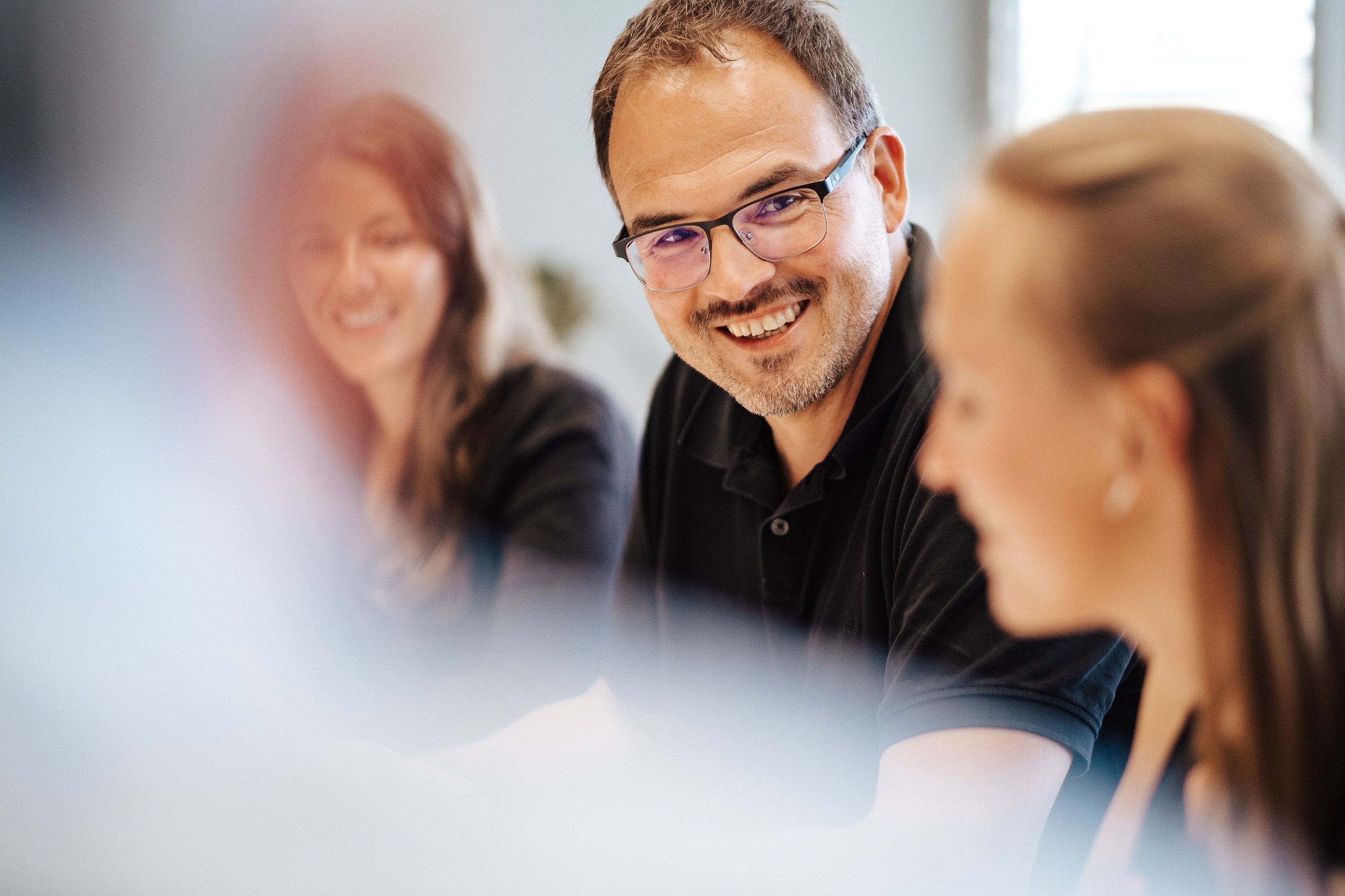 Ein lächelnder Mann mit Brille, umgeben von zwei Frauen, die in einem hellen Raum an einem Tisch sitzen, während eines Gesprächs.