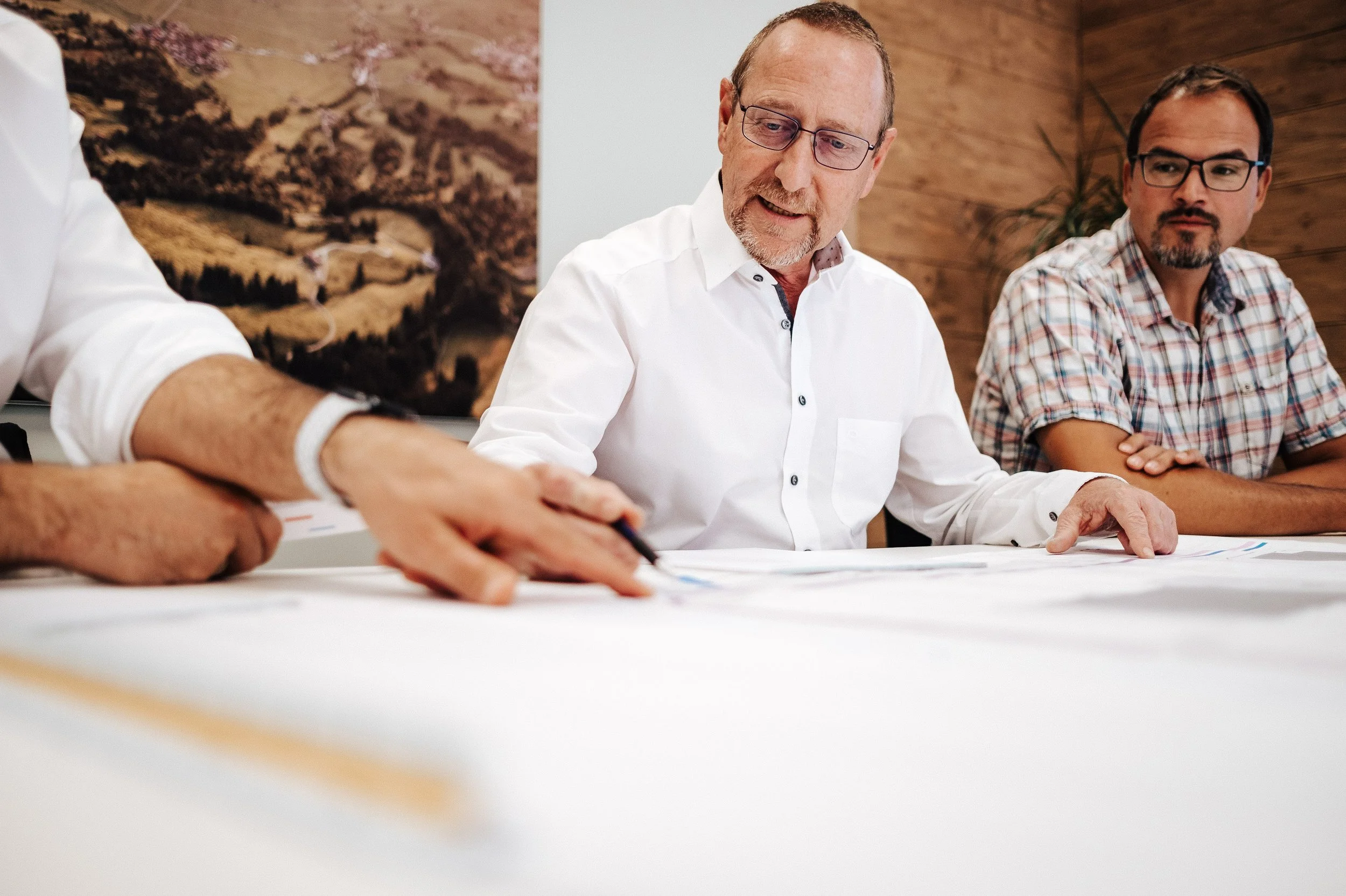 Drei Männer sitzen an einem Tisch, schauen auf Karten oder Pläne, während eines davon mit einem Stift darauf zeigt, in einem modern eingerichteten Raum.