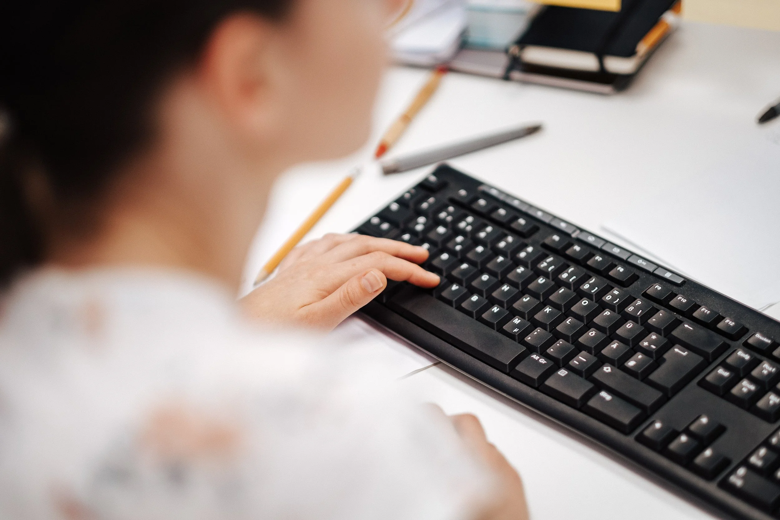 Nahaufnahme einer Person, die an einem Computer arbeitet, Tastatur und Schreibutensilien auf dem Tisch.