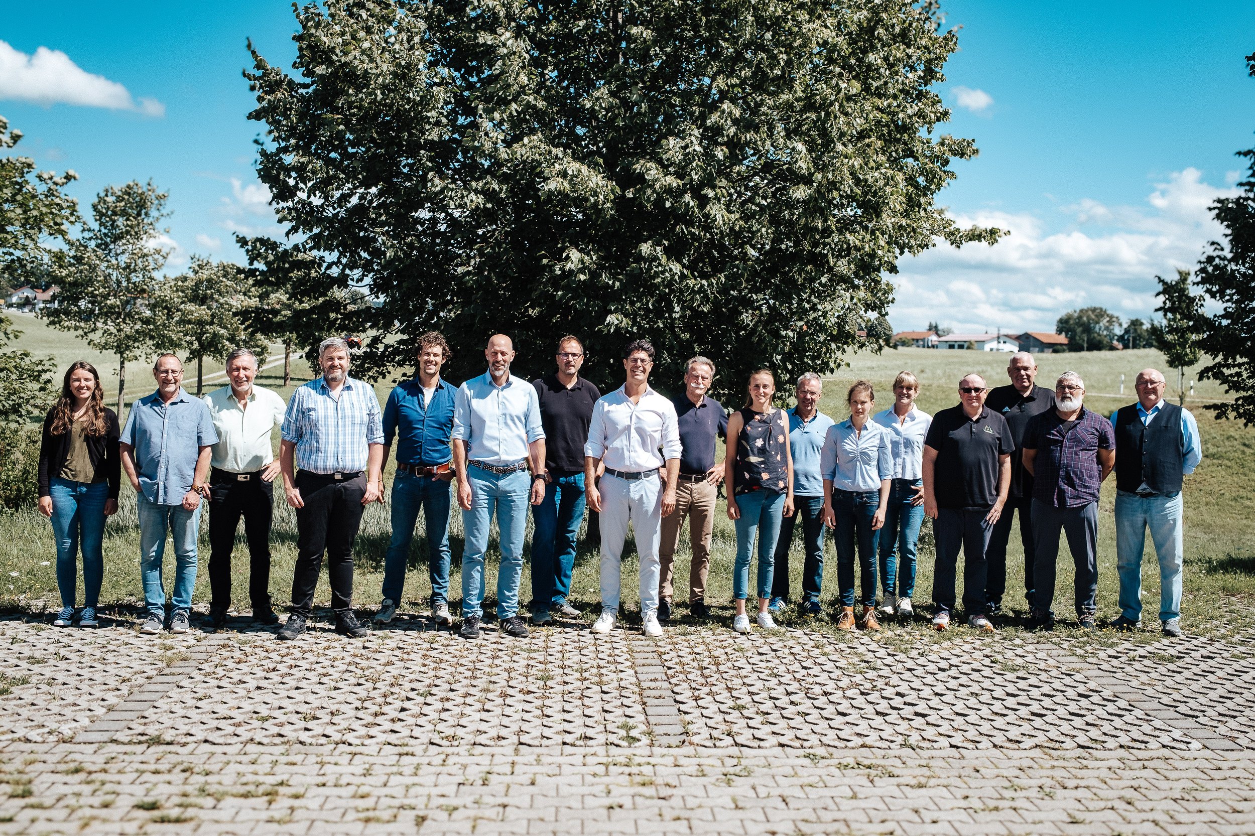 Gruppe von 16 Personen, Männer und Frauen, stehend vor einem großen Baum im Freien bei schönem Wetter.