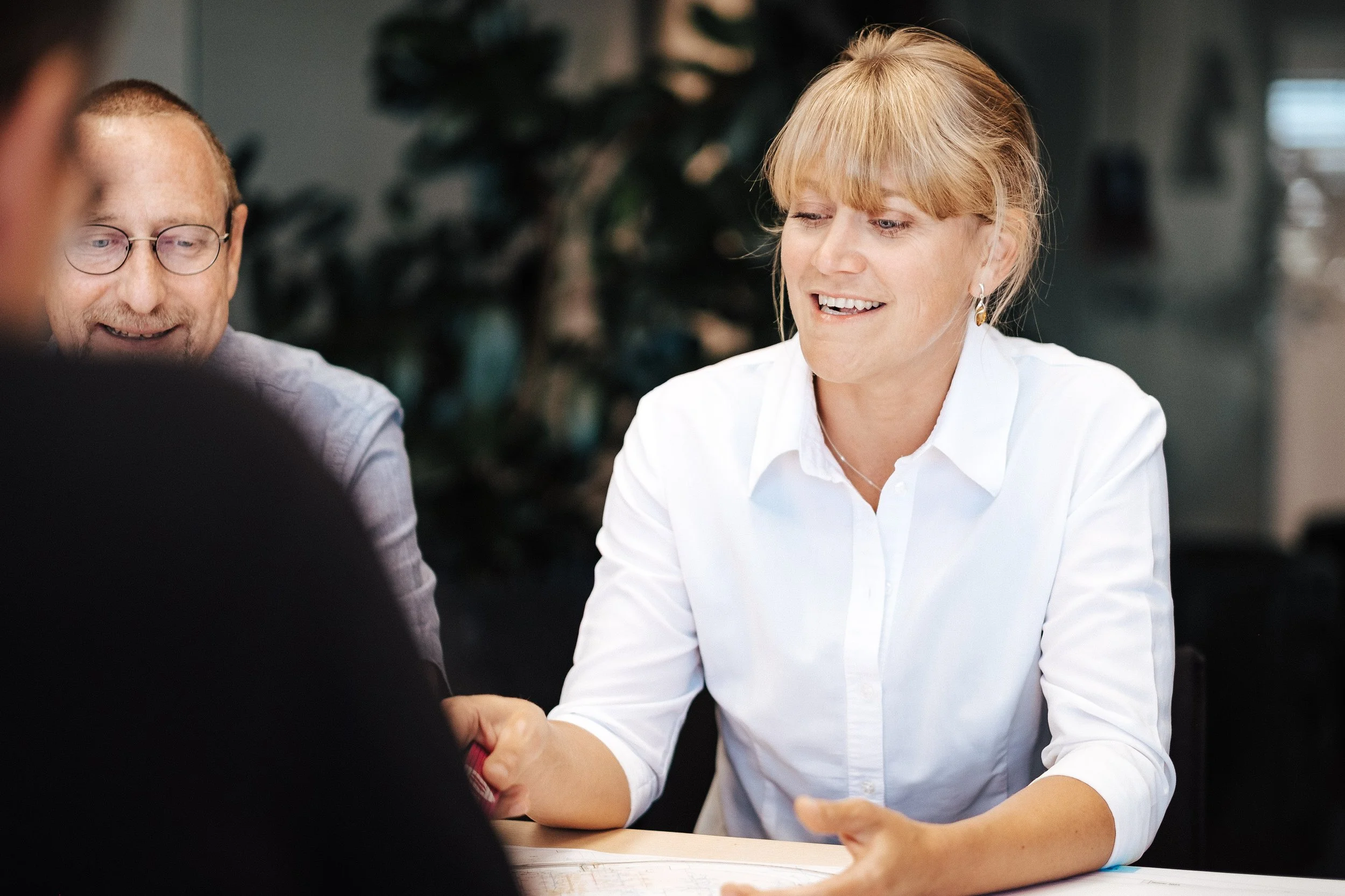 Eine Frau in weißem Hemd schüttelt die Hand eines anderen, während zwei Männer lächelnd zuschauen.