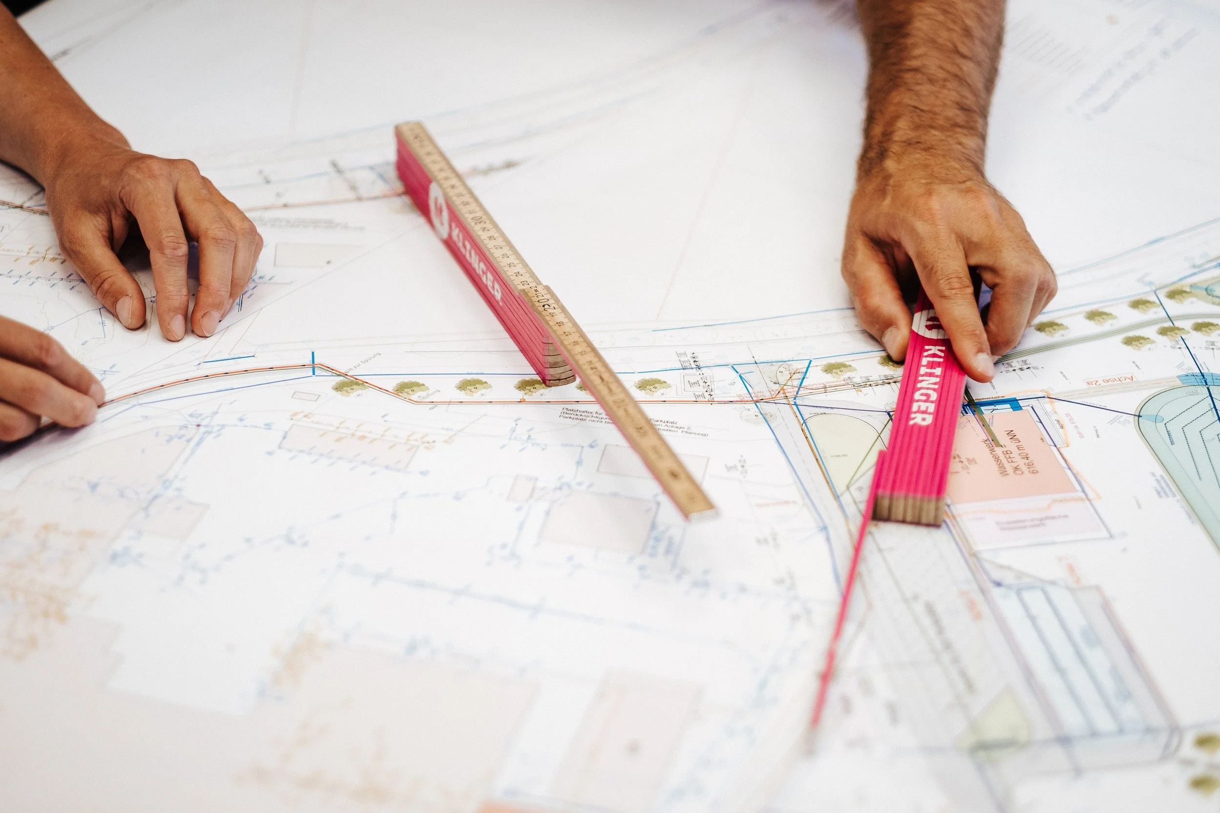 Zwei Hände, die an einem technischen Plan arbeiten, mit einem Lineal und einem kurzen Holzmaßstab.
