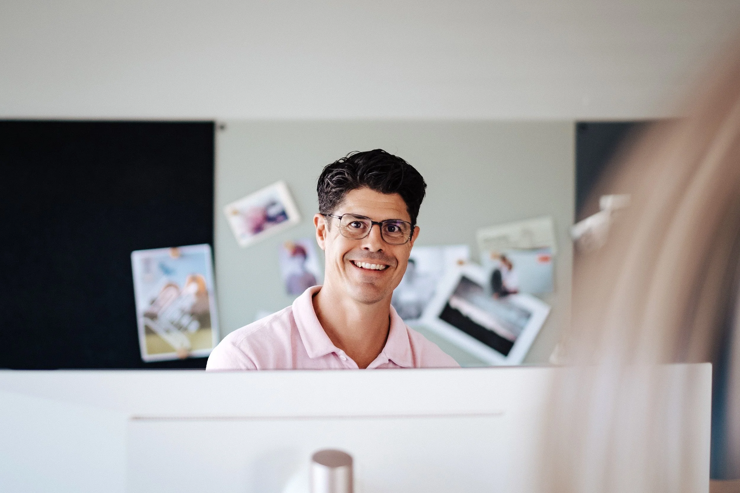 Ein Mann mit dunklem Haar, Brille und rosa Hemd lächelt in die Kamera, im Hintergrund sind Fotos an der Wand.