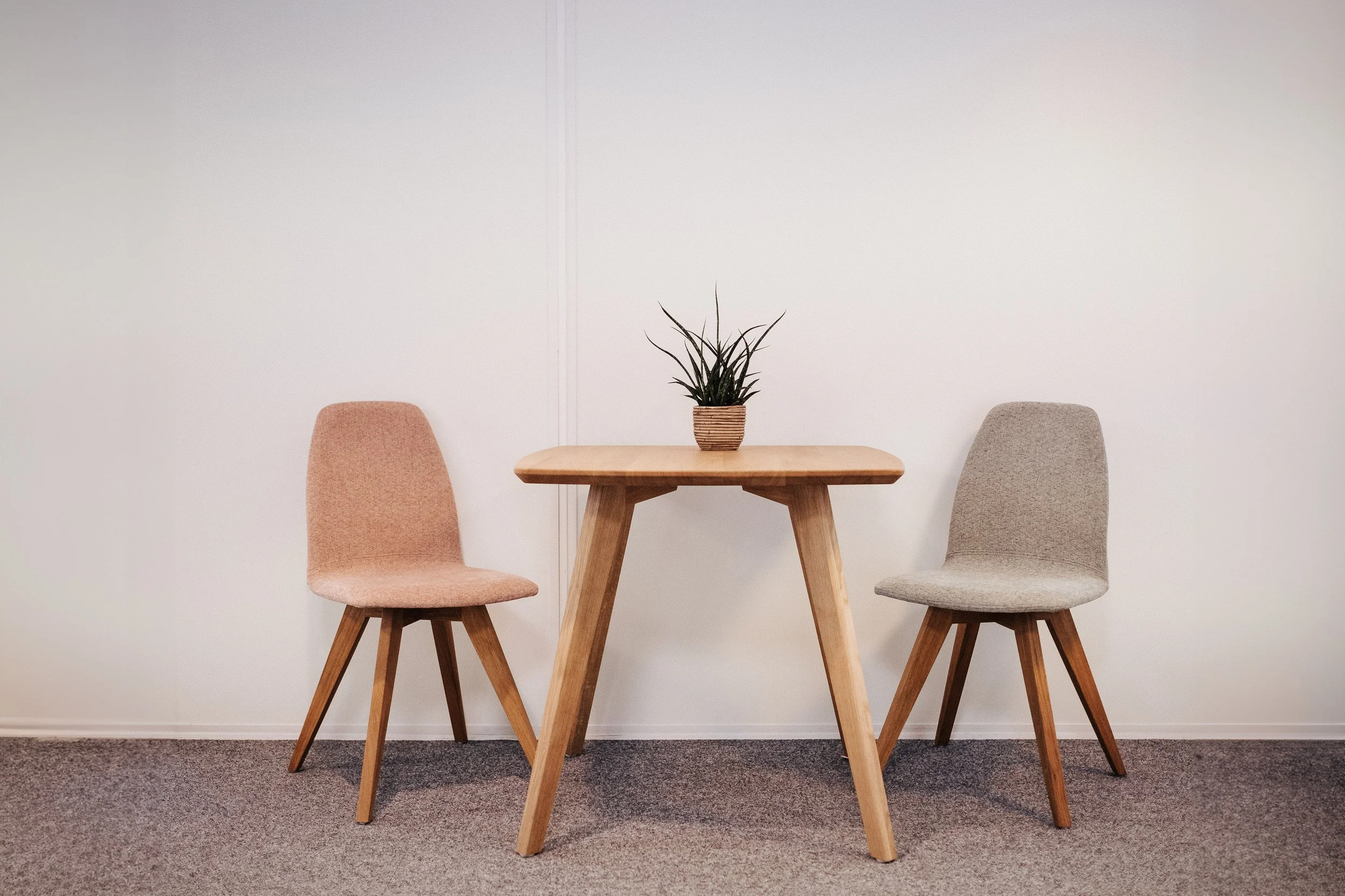 Ein Holztisch mit zwei Stühlen, einer links in hellrosa, einer rechts in beige, mit einer Pflanze in einem Korb in der Mitte auf einem Teppich vor einer weißen Wand.