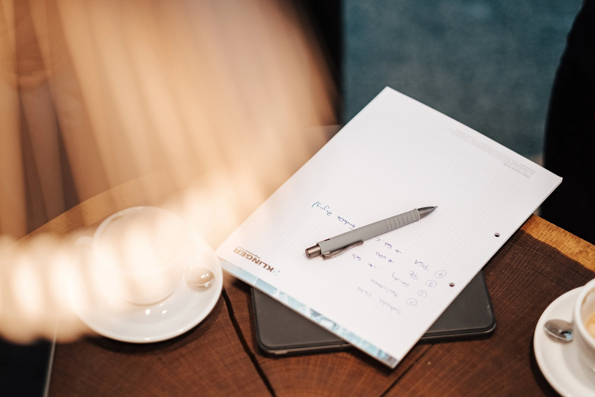Notizblatt mit einem Kugelschreiber und einem Tablet auf einem Holztisch, daneben eine Tasse mit Kaffee.