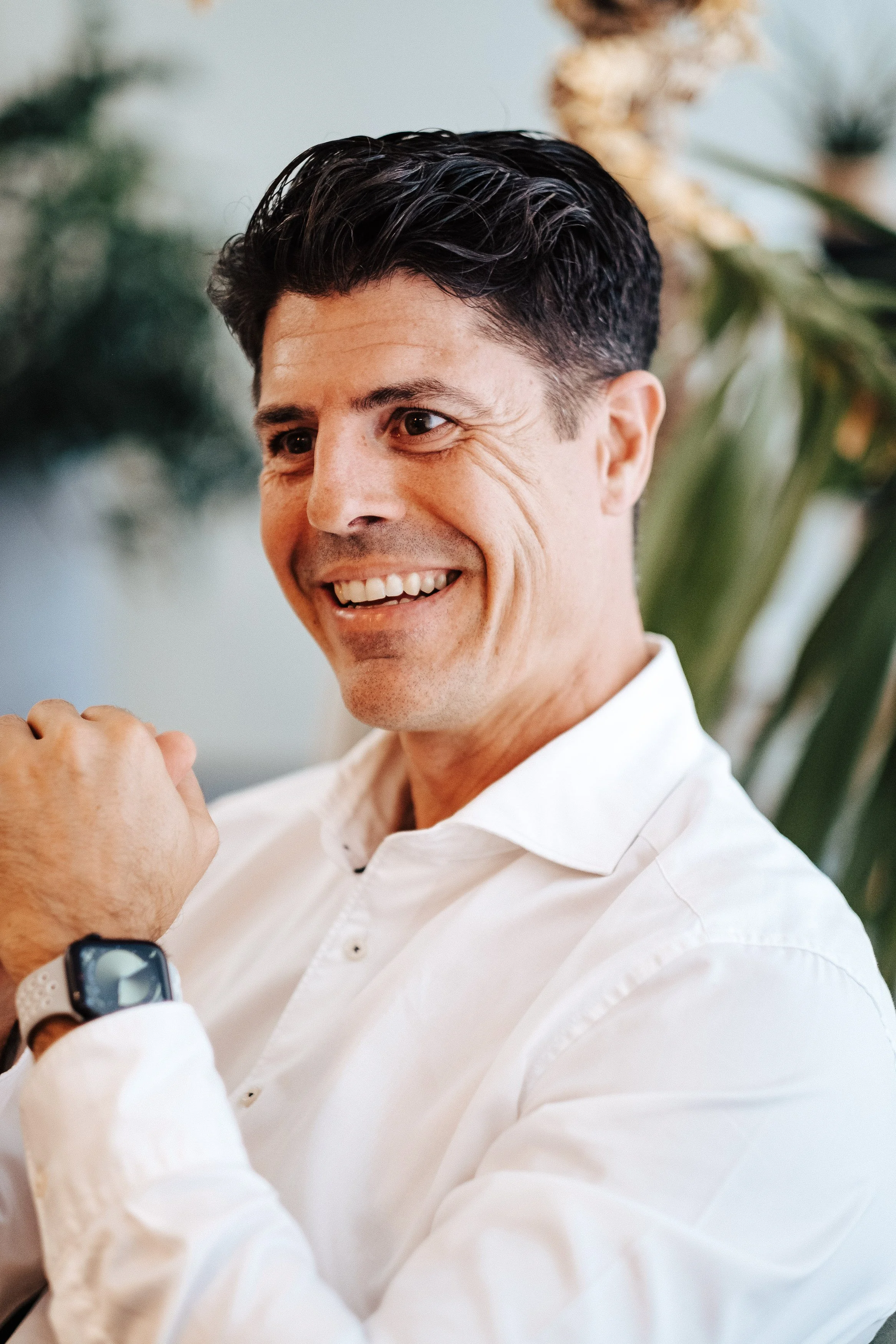 Ein lächelnder Mann in einem weißen Hemd trägt eine Smartwatch und schaut auf seine Uhr, im Hintergrund sind grüne Pflanzen sichtbar.