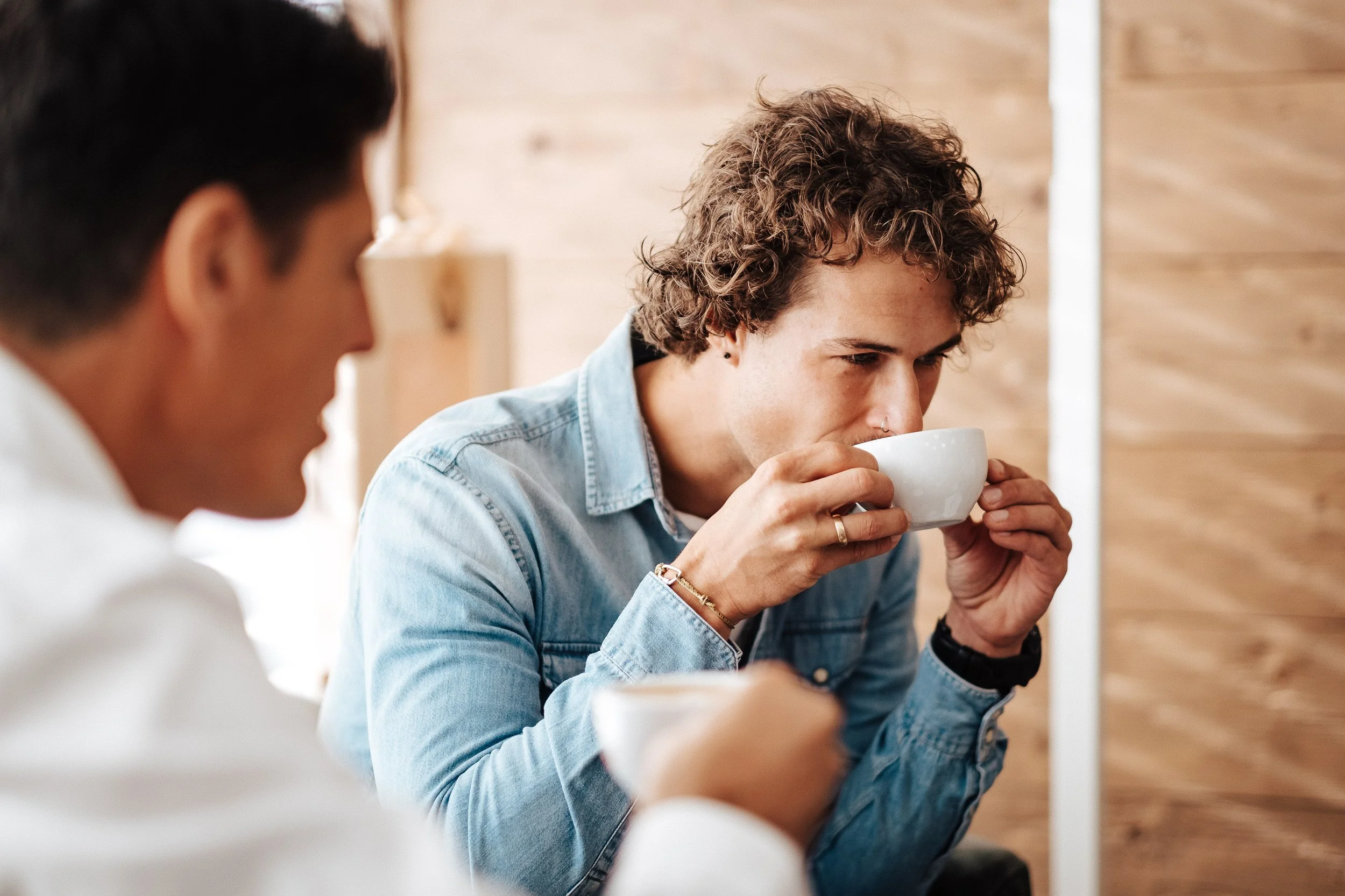 Zwei Männer trinken Kaffee in einem Café, einer trägt eine Jeansjacke und einen Ring, der andere ist unscharf im Vordergrund.