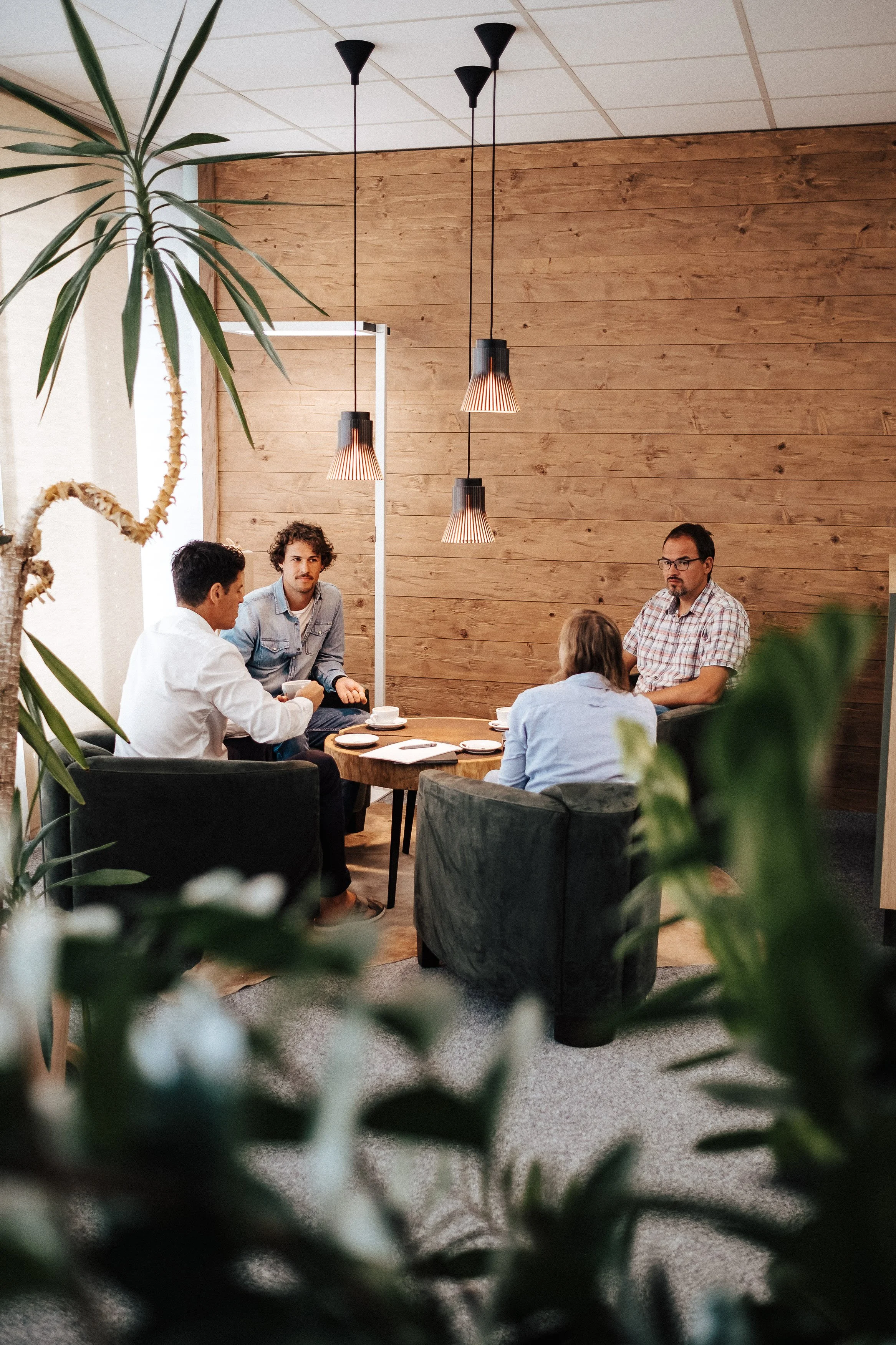 Vier Menschen sitzen in einem modernen Raum mit Holzpaneelen an einem runden Tisch und führen eine Diskussion. Der Raum ist mit Pflanzen, schwarzen Scheinwerfern und einer Decke mit quadratischem Muster dekoriert.