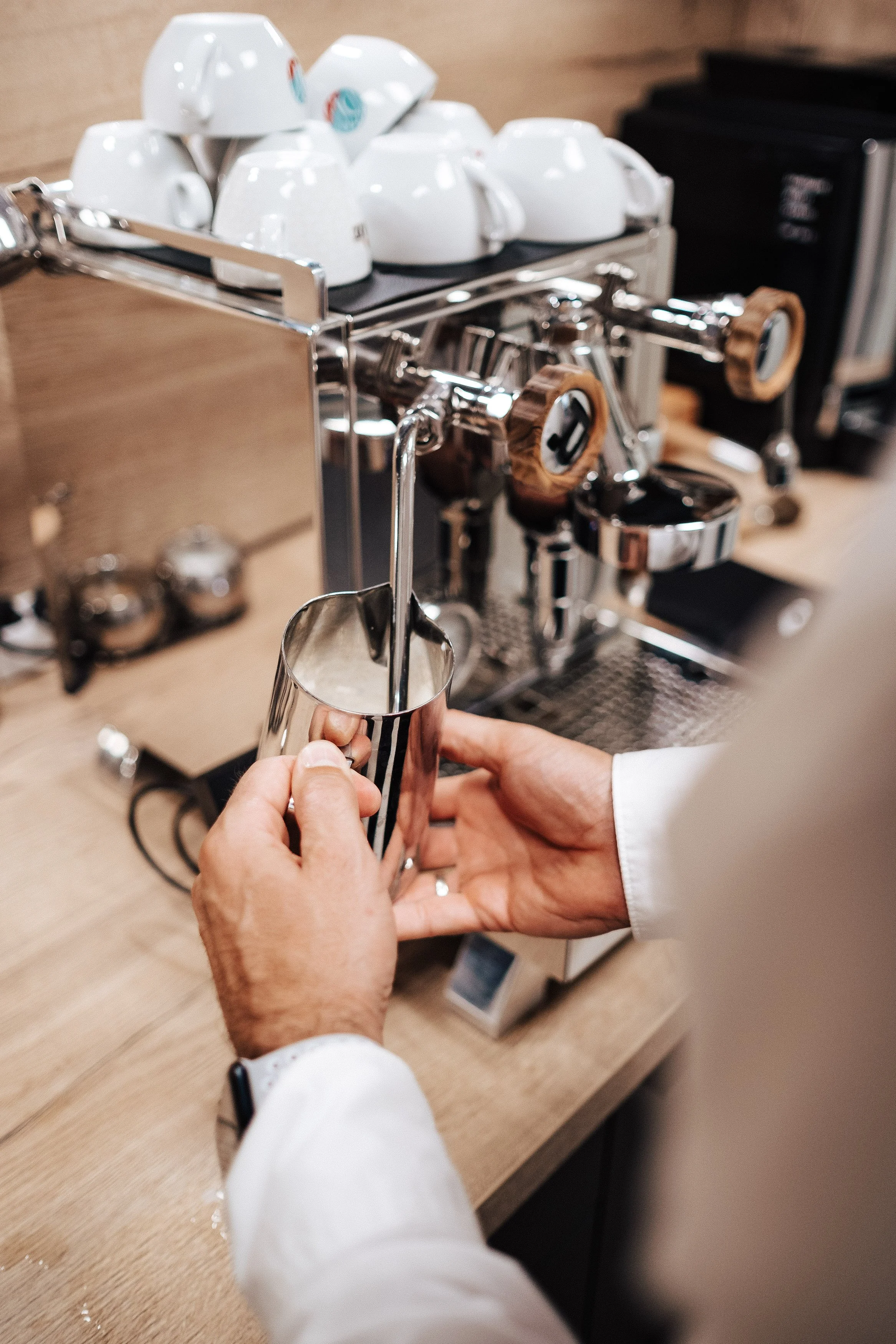 Person macht Milchschaum mit einer Dampflanze an einer Espressomaschine, im Hintergrund Tassen und Kaffeegeräte.