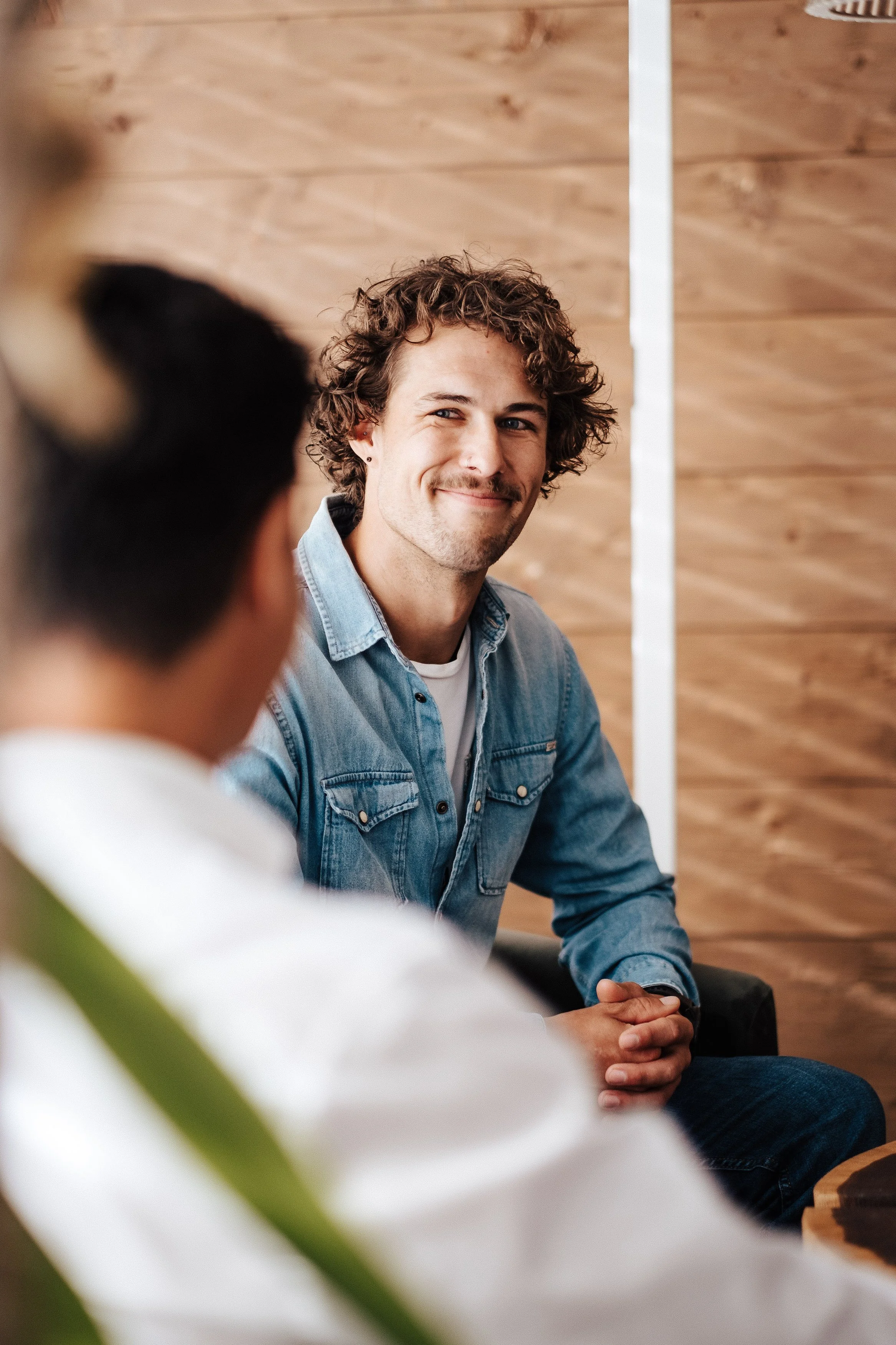 Ein junger Mann mit lockigem Haar und einem leichten Lächeln sitzt an einem Tisch, im Hintergrund eine Holzverkleidung.