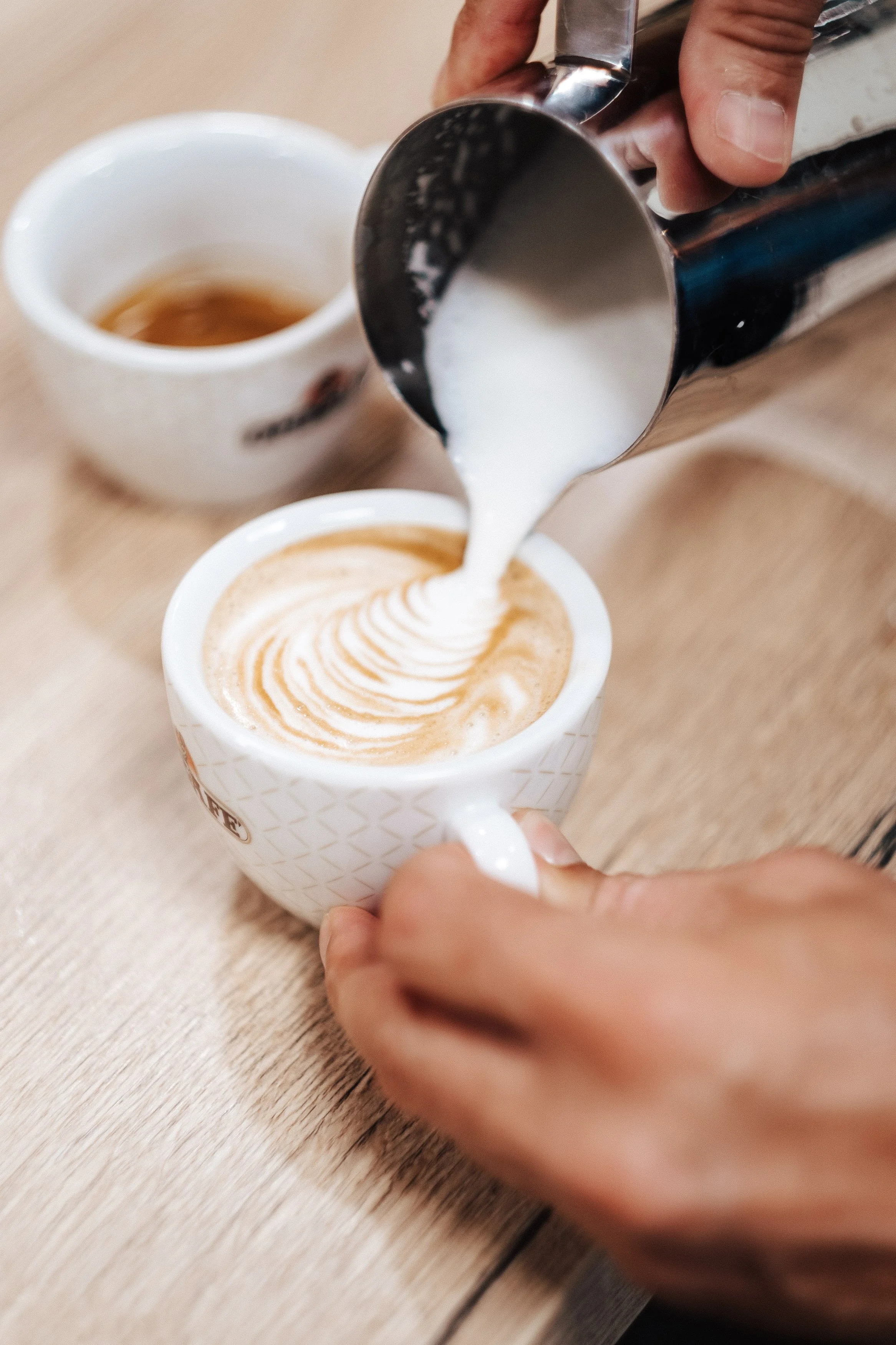 Jemand gießt Milch in einen Cappuccino in einer weißen Tasse auf einem Holztisch, während eine zweite Tasse Kaffee im Hintergrund zu sehen ist.