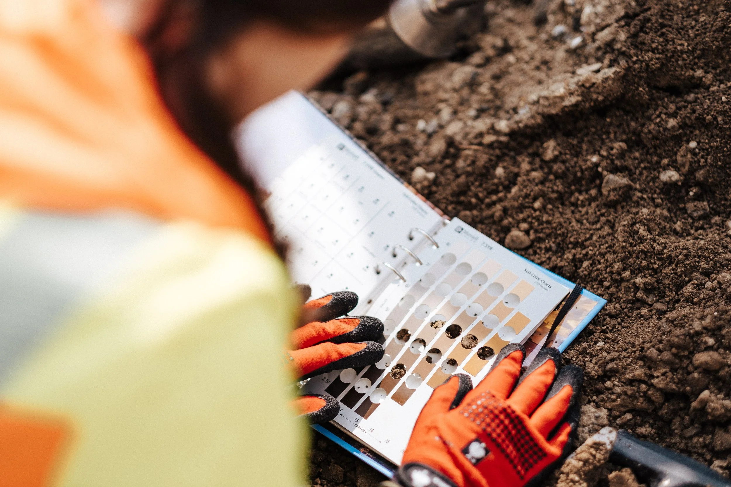Nahaufnahme einer Person mit orangefarbener Schutzjacke und Handschuhen, die einen Boden-Probensammler auf dem Boden neben Erde hält, während sie ein Farbschema in einem Ringbuch betrachtet.