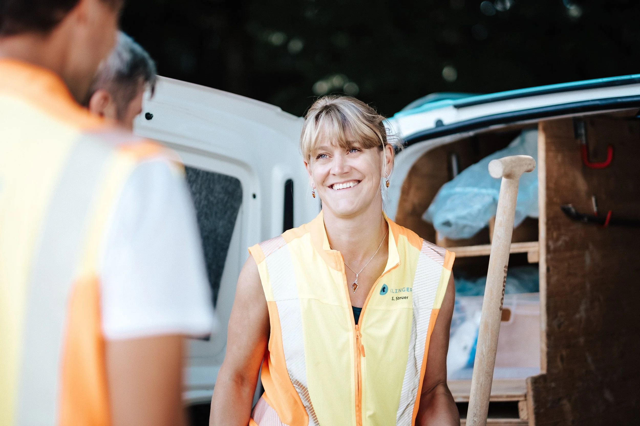 Eine Frau mit einem gelben Sicherheitsweste steht neben einem weißen Transportfahrzeug, während sie mit anderen Personen spricht, im Hintergrund sind Baum und Arbeitsmaterialien
