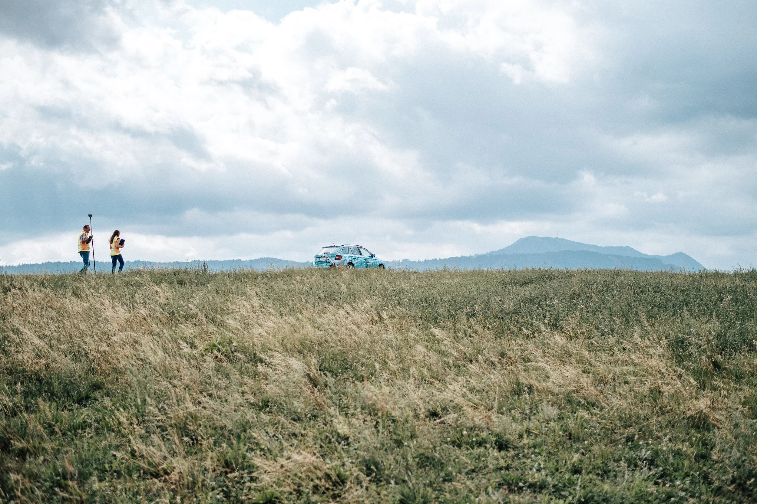 Zwei Personen mit Messgerät und Notizblock auf einer Wiese, in der Ferne ein Auto und Berge bei bewölktem Himmel.