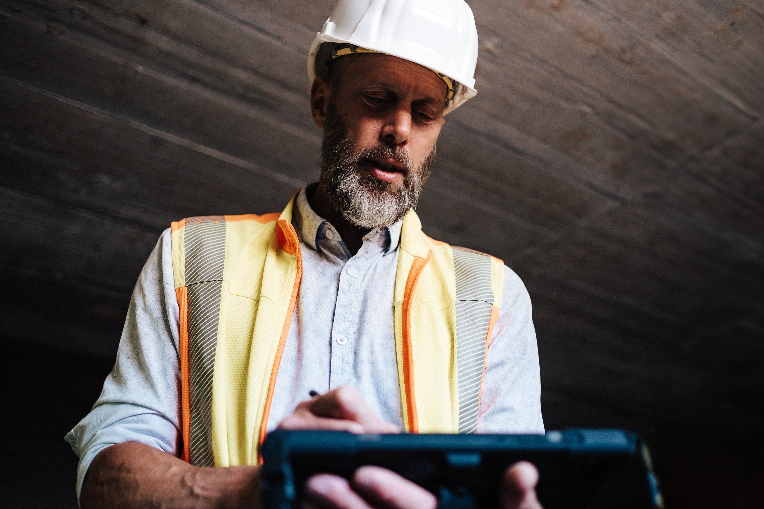 Ein Mann trägt einen weißen Schutzhelm und eine reflektierende Warnweste, während er auf einem Baugrundstück oder in einer Baustelle arbeitet und eine Schreibtafel in der Hand hält.