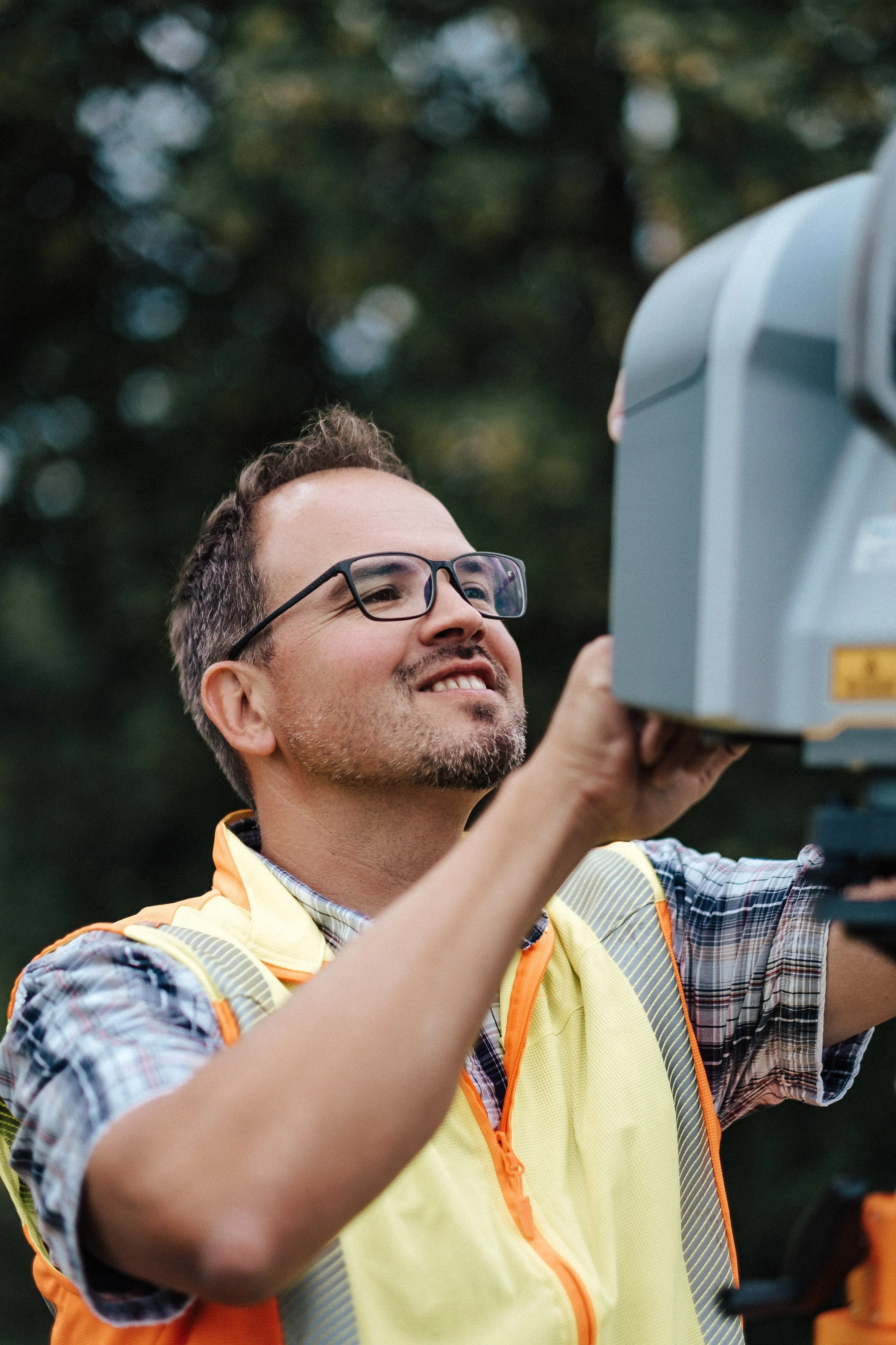 Ein Mann mit Brille und Bart, trägt eine gelbe Sicherheitsweste und prüft mit einem Messgerät im Freien, im Hintergrund sind Bäume sichtbar.