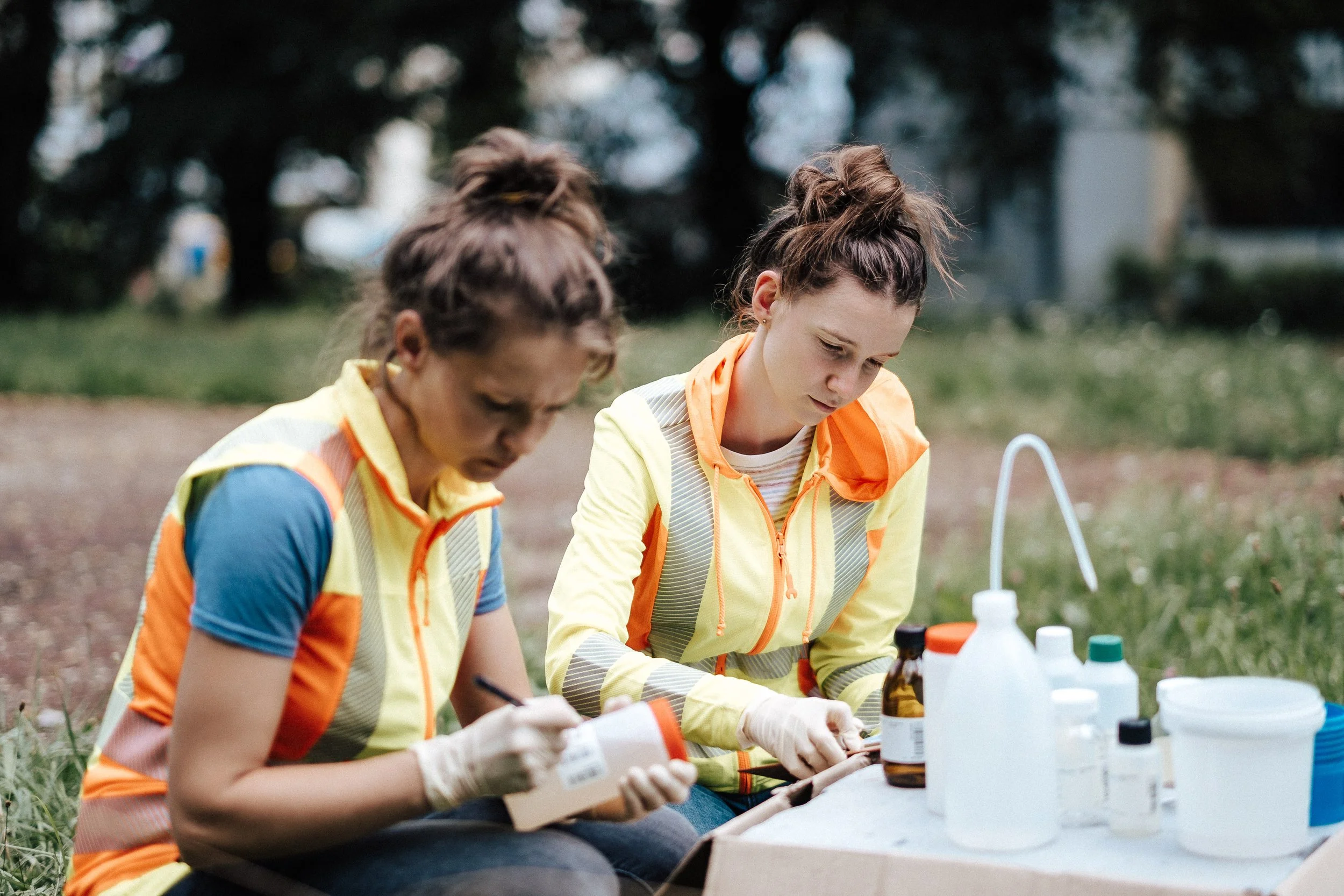 Zwei junge Frauen in Sicherheitskleidung, die draußen am Tisch mit Medikamenten und Laborutensilien arbeiten.