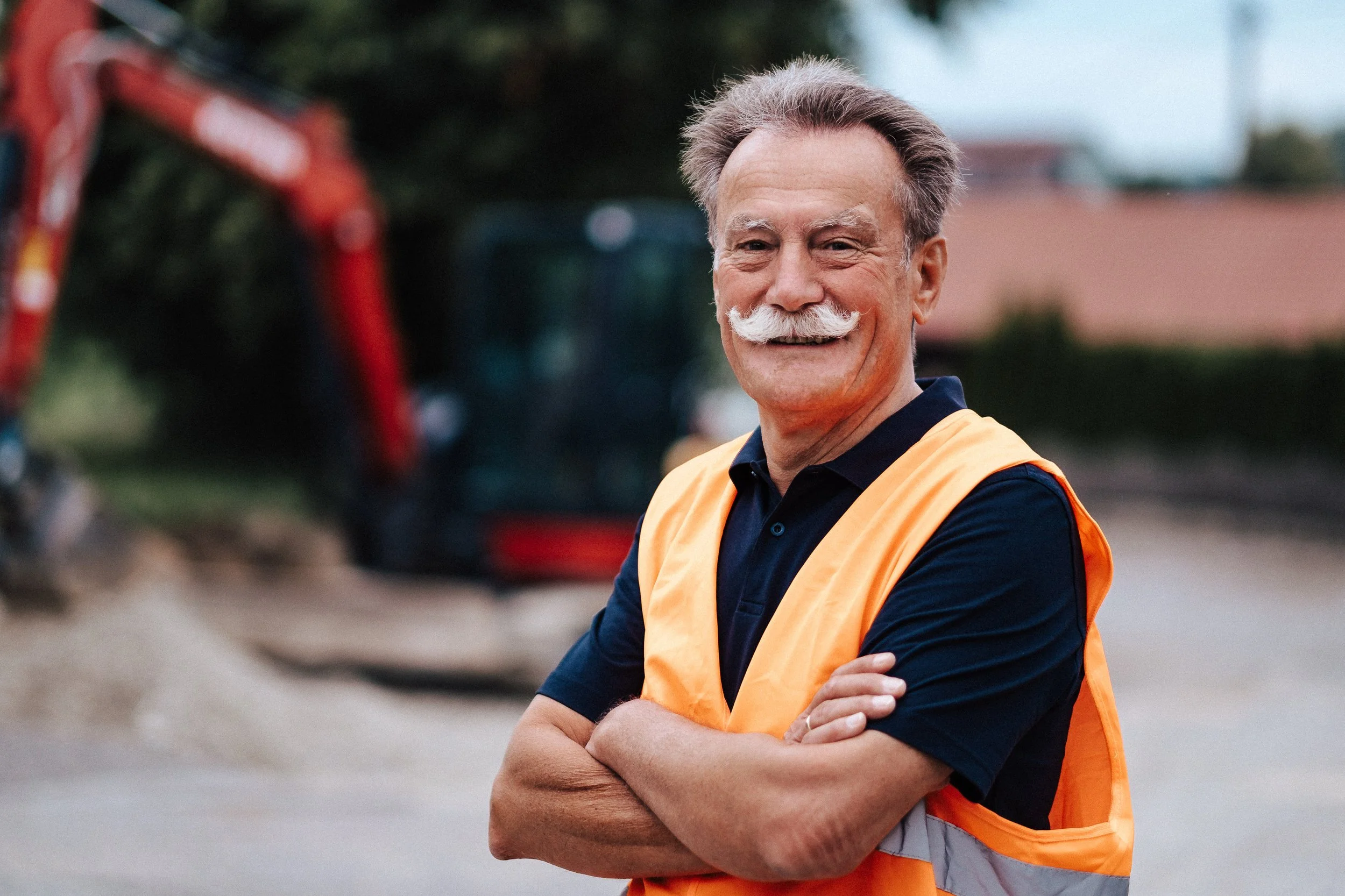 Ein älterer Mann mit grauem Haar und Schnurrbart trägt eine dunkelblaue Poloshirt und eine orange Sicherheitsweste. Er steht mit verschränkten Armen vor einer Baustelle, im Hintergrund sind Baumaschinen und eine Gebäudestruktur zu sehen.