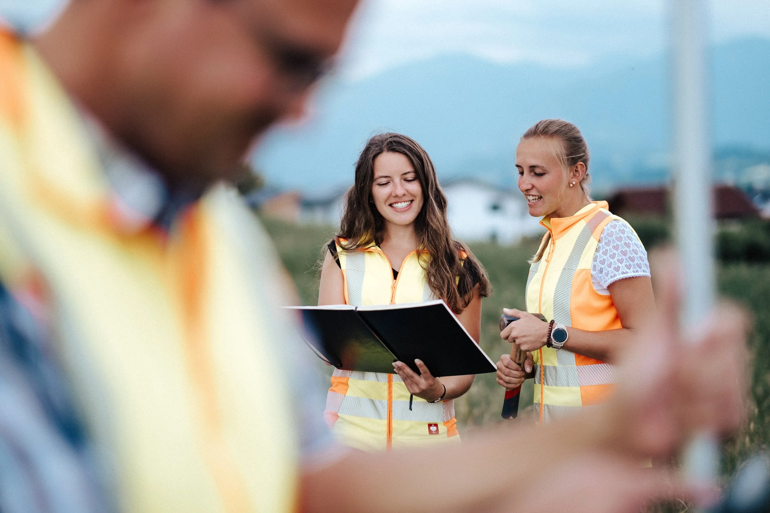 Drei Bauarbeiterinnen mit reflektierenden Sicherheitswesten, die draußen auf einer Baustelle stehen und sich unterhalten, während die Frau in der Mitte eine Mappe hält und lächelt.