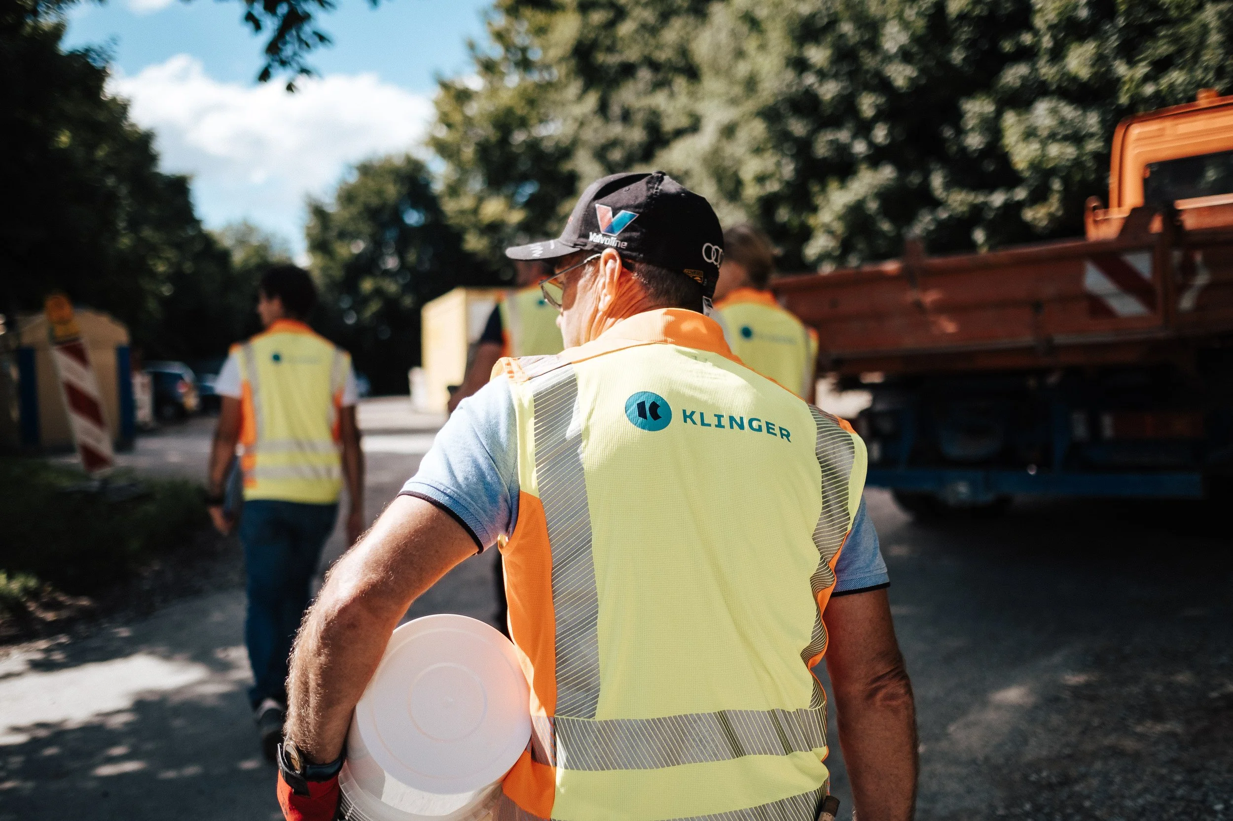 Menschen in Warnwesten unterwegs auf einer Straße, einige tragen Kappen und Sonnenbrillen, im Hintergrund Baum und LKW, sonniges Wetter.