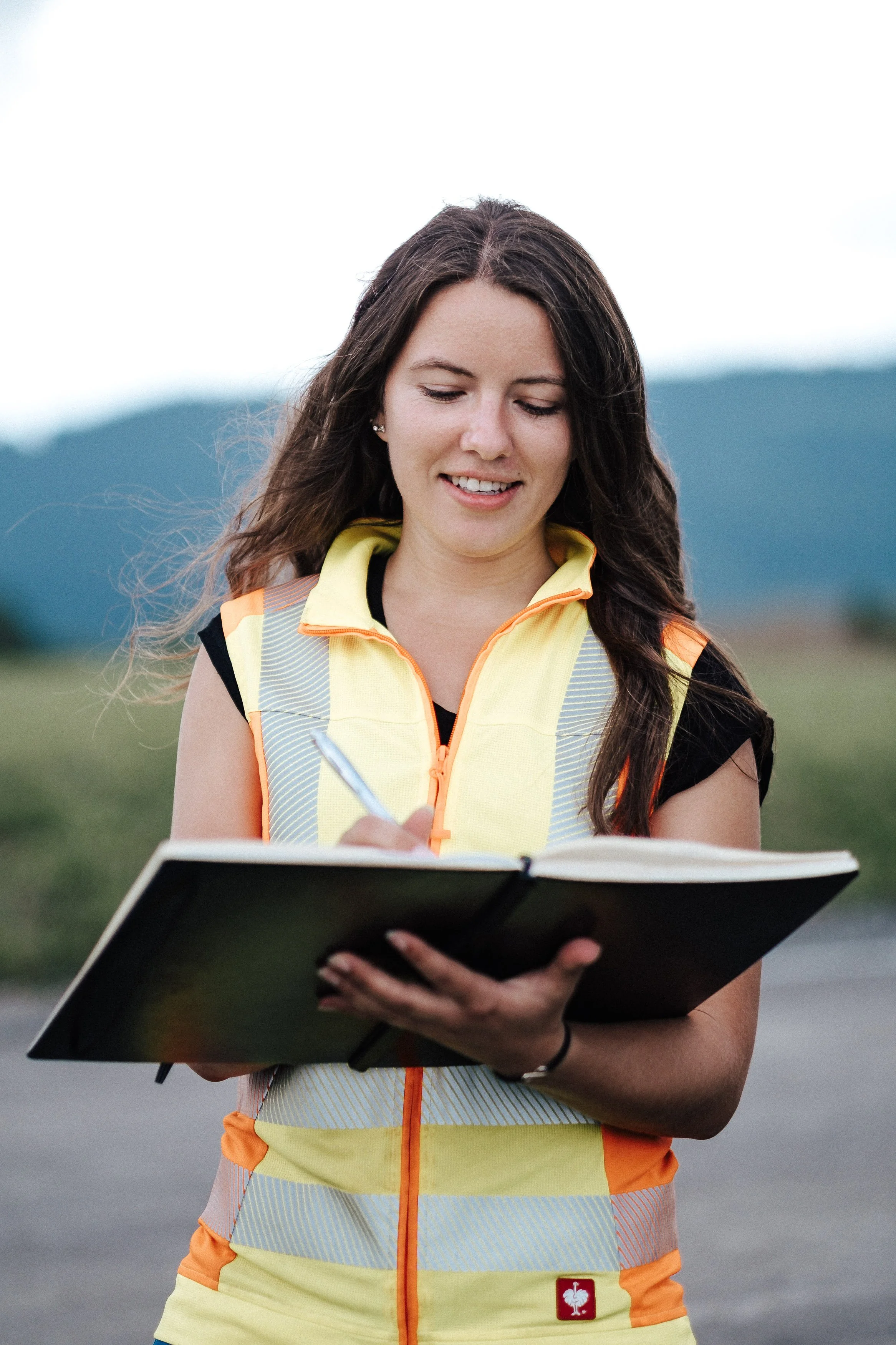 Eine junge Frau trägt eine Sicherheitsweste und schreibt in ein Notizbuch im Freien, mit Bergen im Hintergrund.