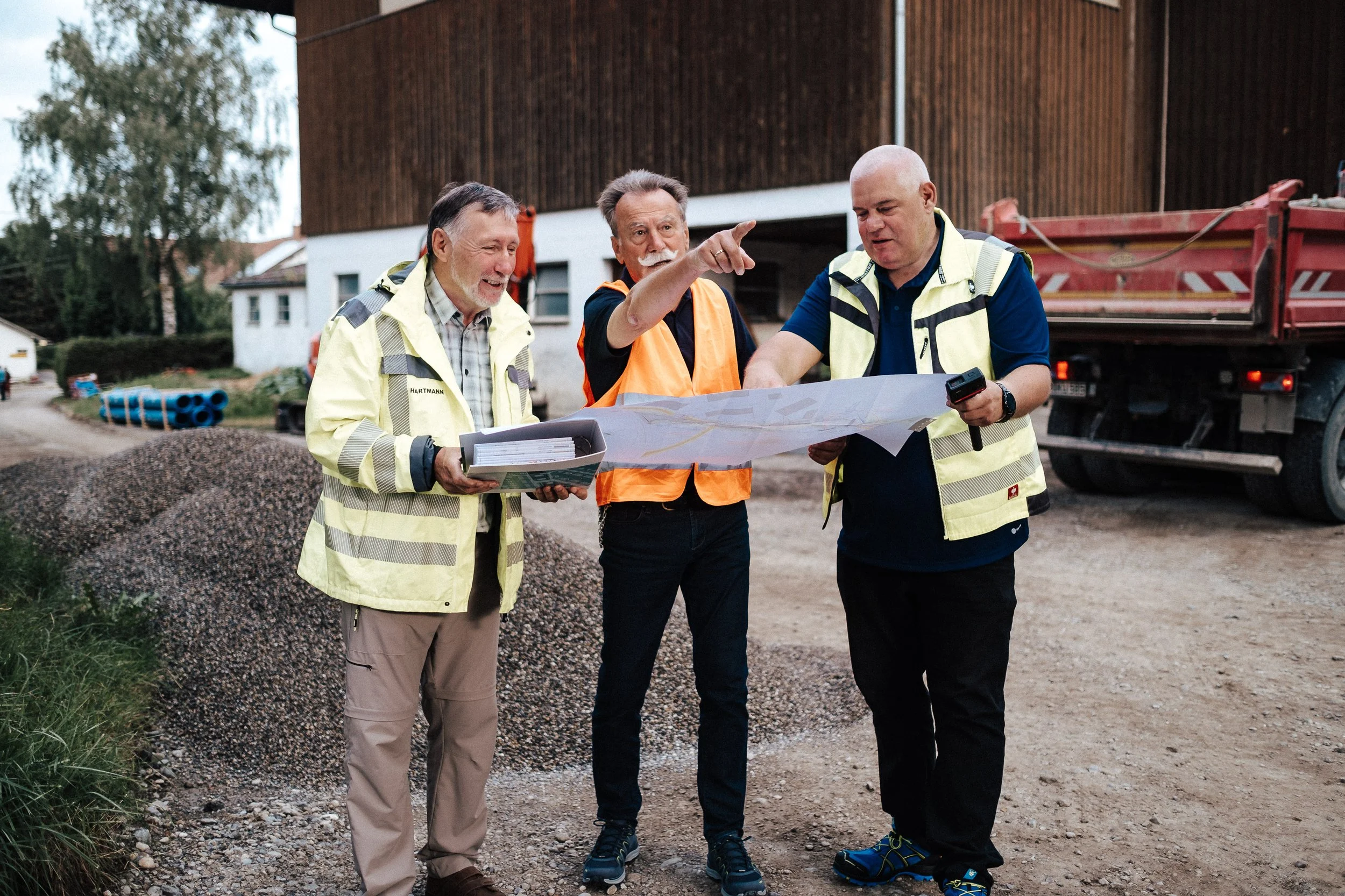 Drei Bauarbeiter schauen auf eine große Karte auf einer Baustelle, mit einem LKW im Hintergrund.