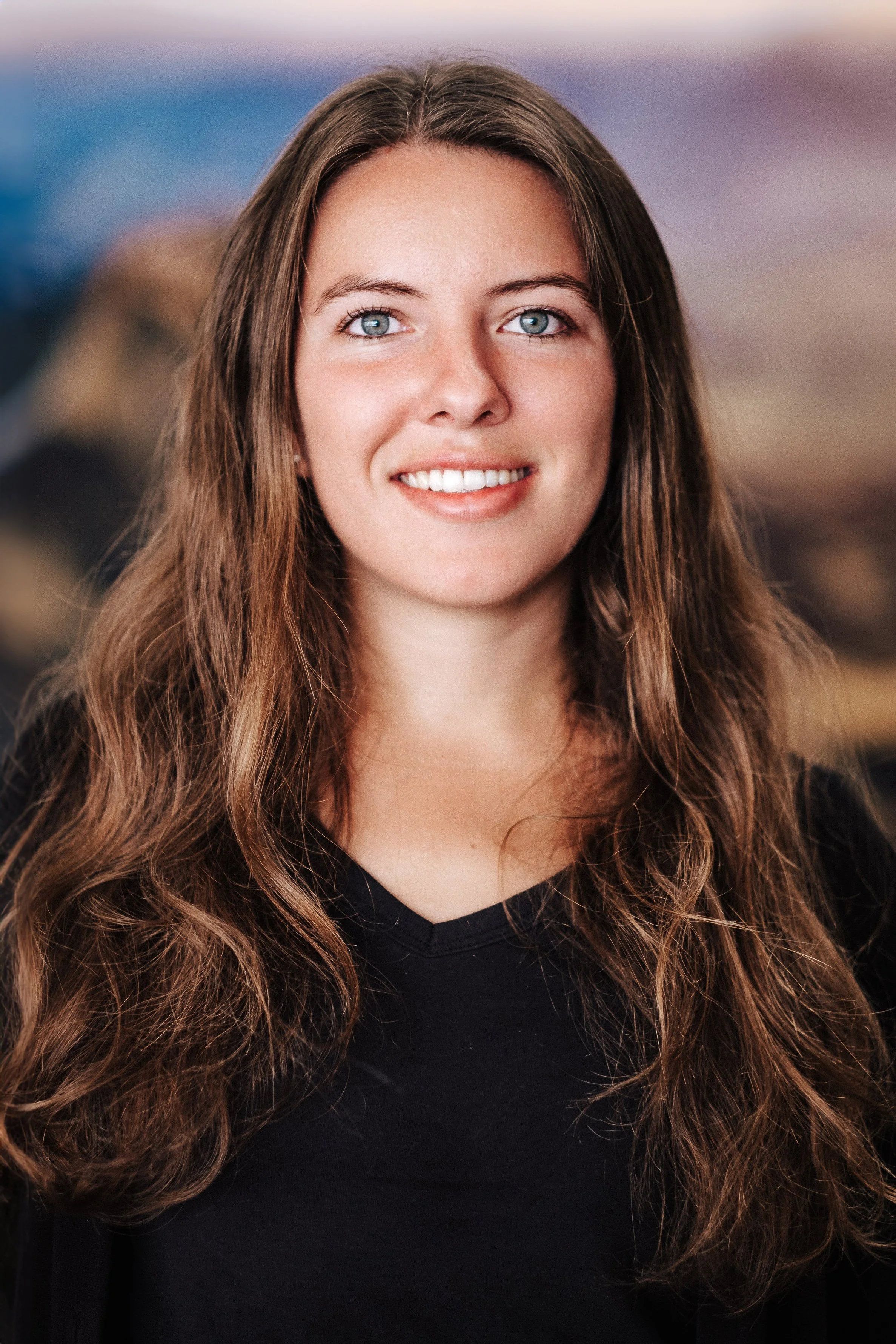 Porträt einer jungen Frau mit langen, lockigen braunen Haaren und blauen Augen, lächelnd, vor einer unscharfen Naturkulisse bei Sonnenuntergang.