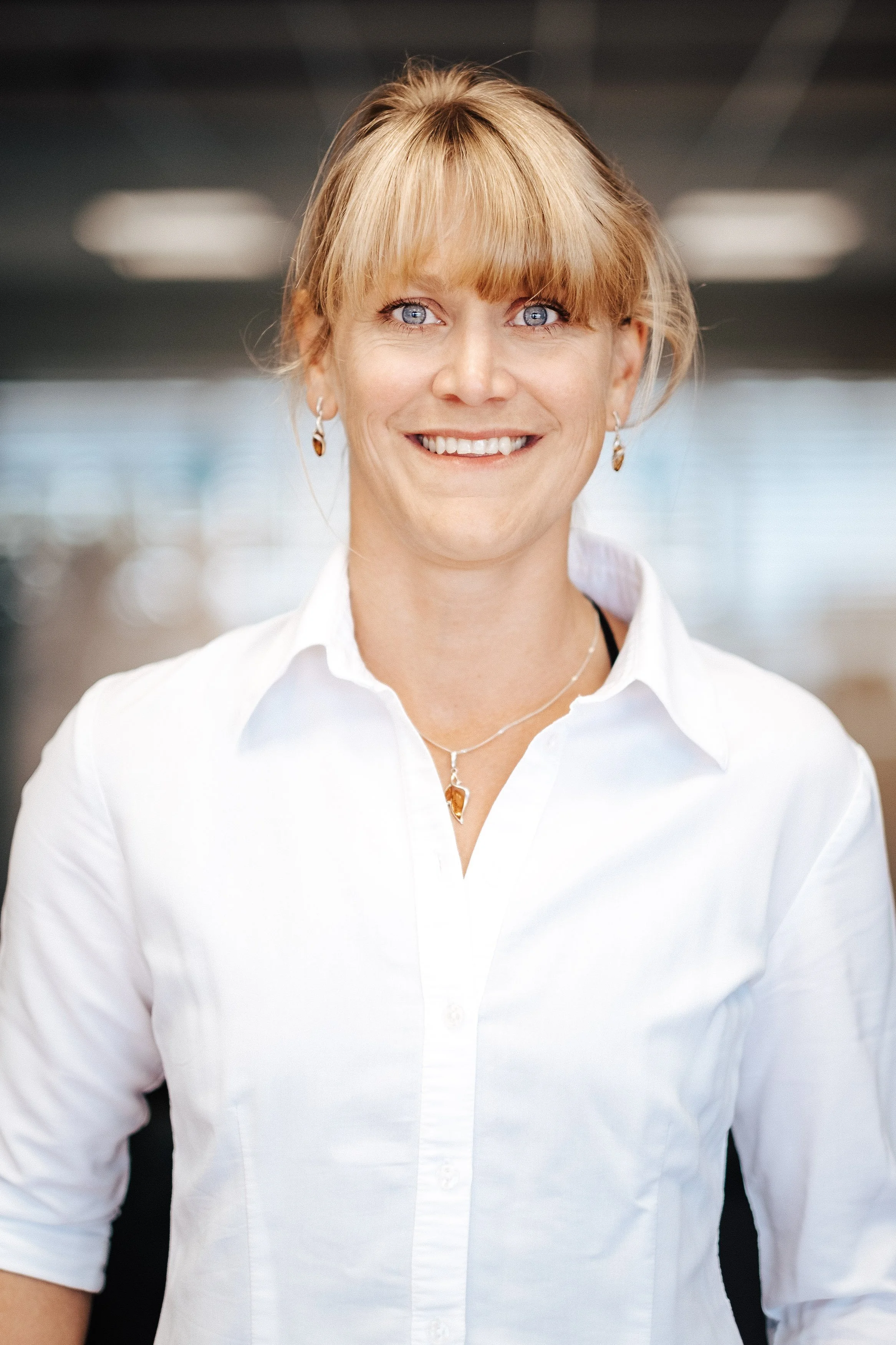 Eine Frau trägt weiße Bluse, hat blonde Haare, trägt Ohrhänger und eine Halskette, lächelt in einem öffentlichen Gebäude mit modernem Design.